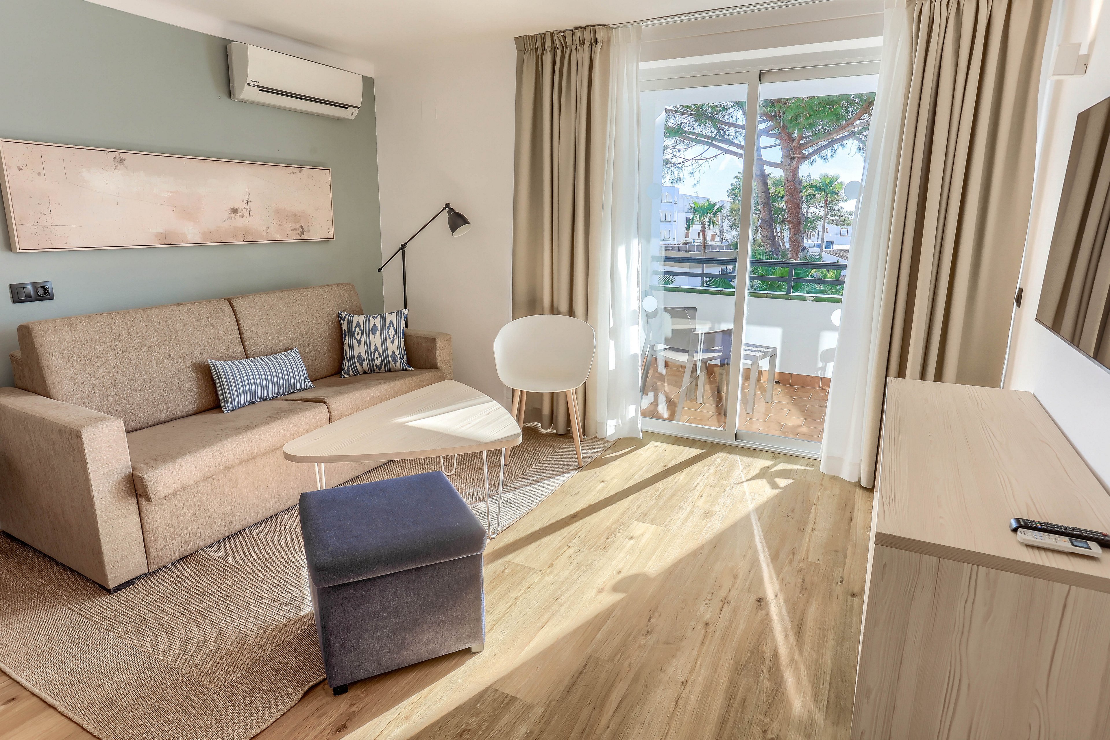A contemporary living room features a beige sofa, accent pillows, and a minimalist coffee table. Large glass doors open to a balcony with outdoor seating, allowing sunlight to fill the space. The room includes wood flooring, neutral tones, and a wall-mounted air conditioner. The setting is calm and inviting, with a mix of modern and cozy elements.