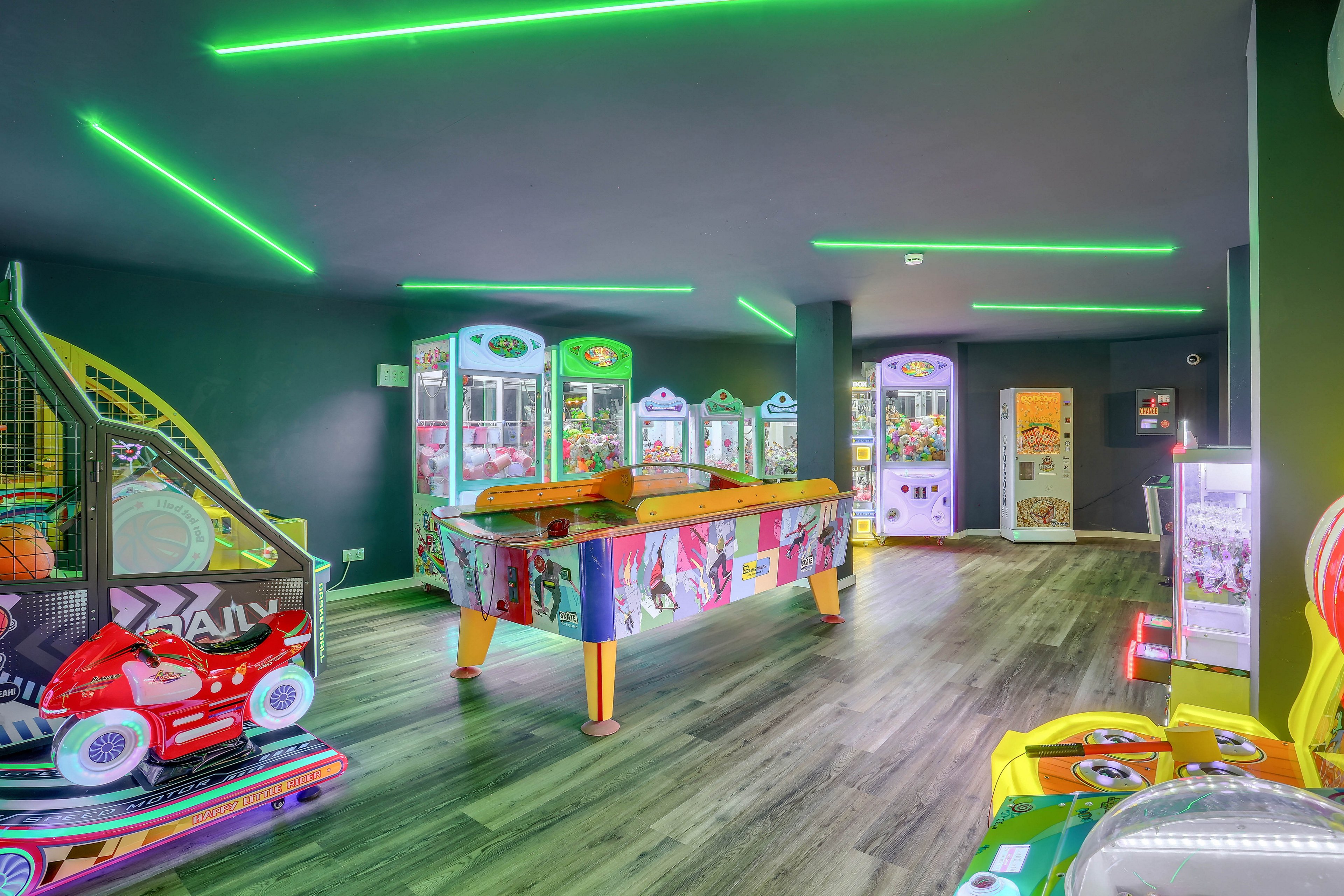 A vibrant arcade room features multiple game machines, including air hockey and claw machines, under striking neon green ceiling lights. The space has a contemporary design with wood flooring and colorful visuals on the game units. No visible text or numeric values are present on the machines. The setting is empty, emphasizing the playful and energetic atmosphere.