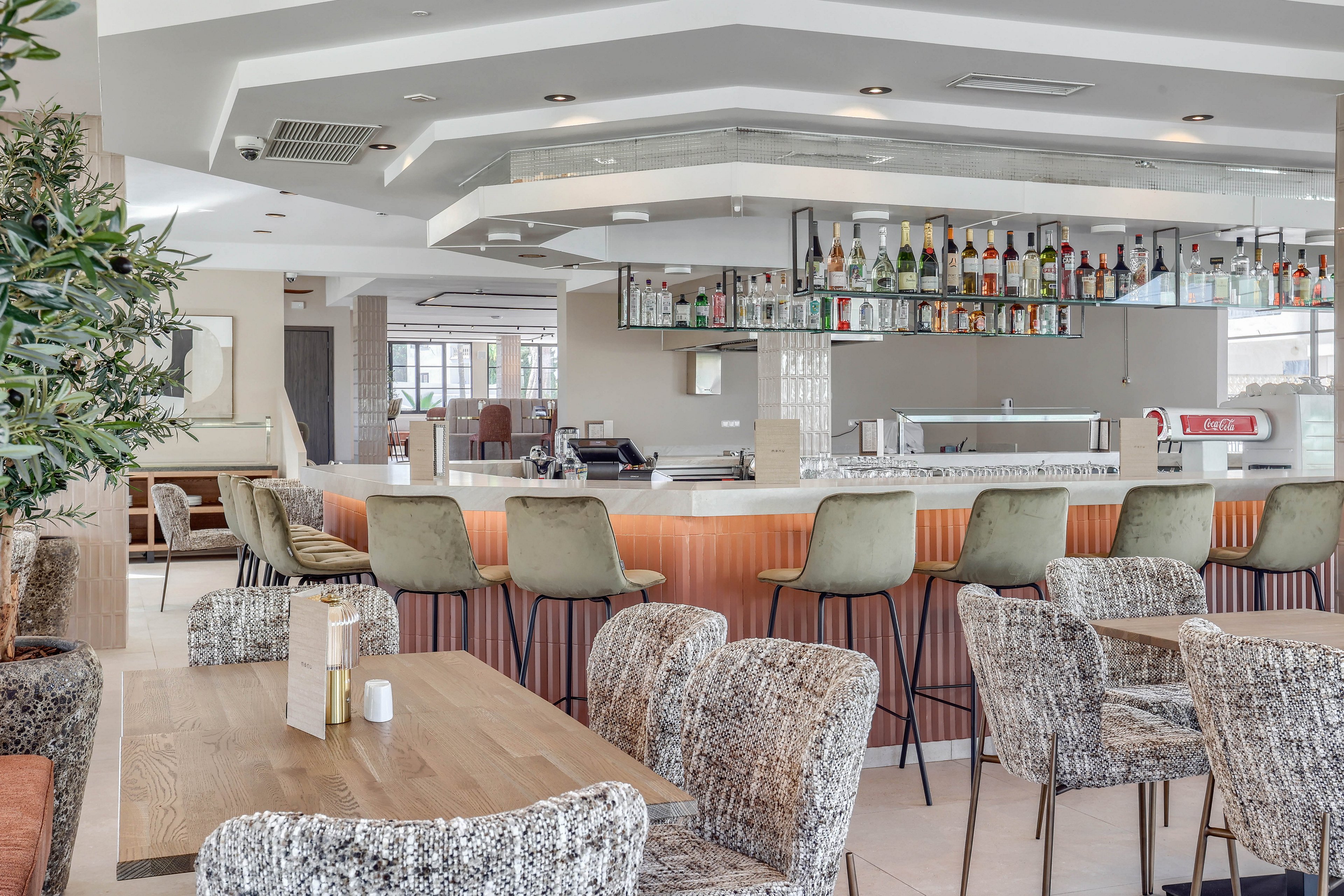 A contemporary hotel bar features a sleek counter with green barstools and a variety of bottles displayed above. The setting is bright and airy, with patterned upholstered chairs and wooden tables arranged for casual dining. The neutral color palette and clean lines create a welcoming, modern atmosphere. No people are present, emphasizing the inviting and organized space.