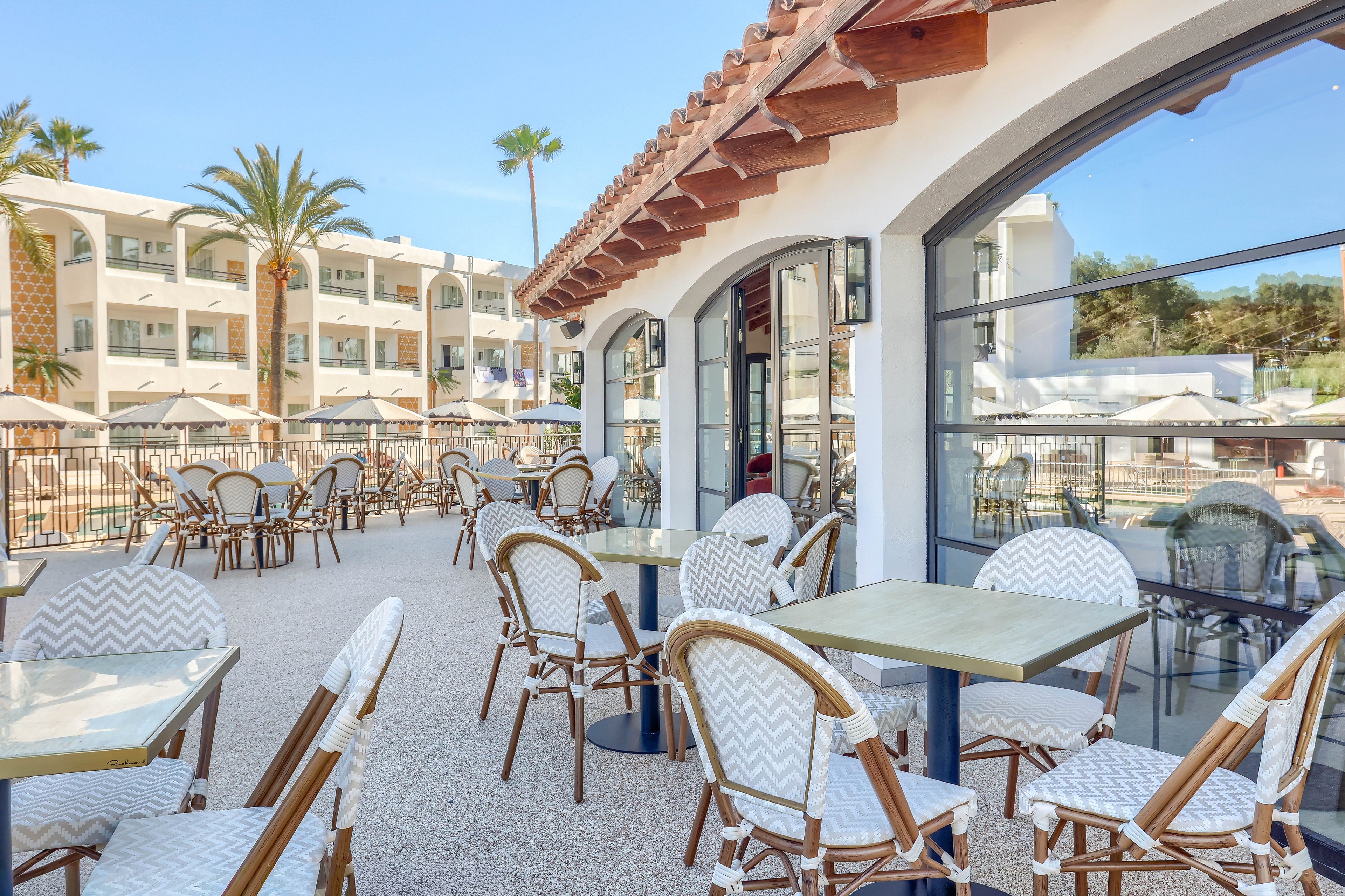 A spacious outdoor terrace features several empty tables and chairs arranged for dining. The setting is adjacent to a modern hotel or resort building with white walls and large windows, surrounded by palm trees. The area is brightly lit with natural sunlight, creating a relaxed and inviting atmosphere. No people are present, and the scene highlights the clean, contemporary design and vacation ambiance.