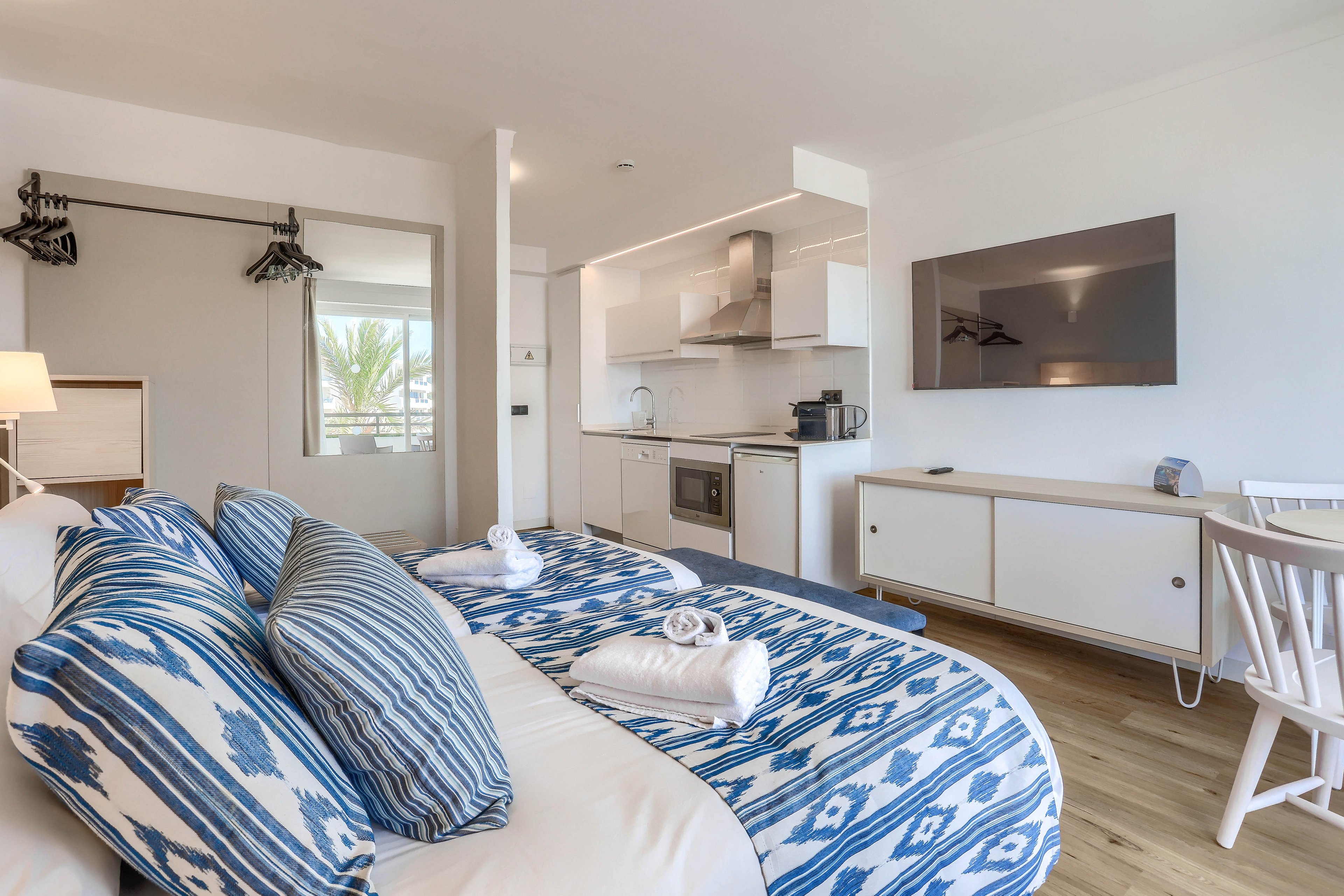A contemporary studio apartment features a double bed with blue and white patterned bedding and neatly folded towels. The space includes a kitchenette with appliances, a large wall-mounted television, and a white desk with a chair. Natural light fills the room, highlighting the clean, minimalist decor and wooden flooring.