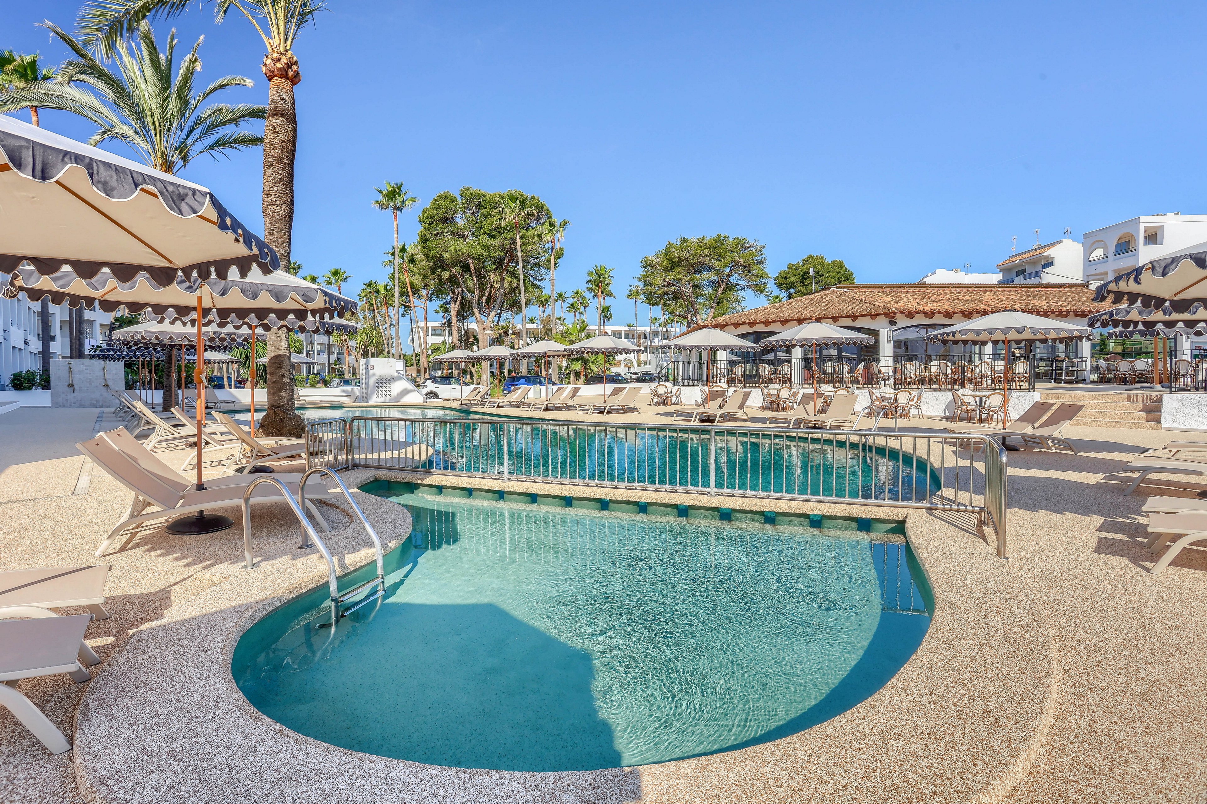The image features a tranquil outdoor swimming pool surrounded by numerous lounge chairs and shaded umbrellas. The setting is a resort or hotel, with palm trees and white buildings visible in the background. The scene is bright and inviting, with clear blue skies and a relaxed atmosphere. No people are present, emphasizing the peaceful mood and clean, modern design.
