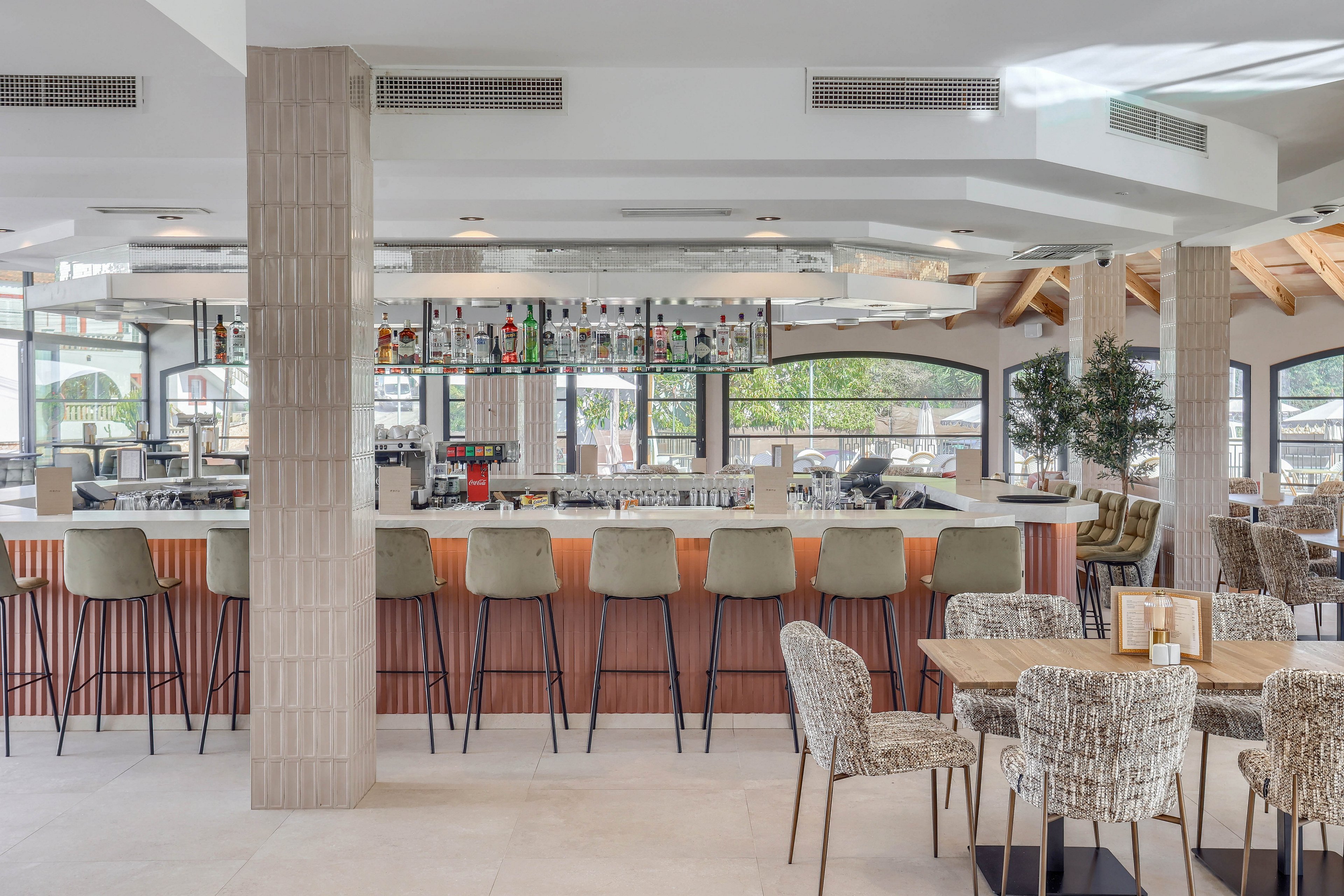 The image features a contemporary bar area with high stools arranged along a wooden counter. The setting is bright and airy, with large windows letting in natural light and neutral tones dominating the palette. The space includes textured chairs and tables, with visible bottles and glassware displayed above the bar. The overall mood is inviting and sophisticated, ideal for social gatherings or casual dining.
