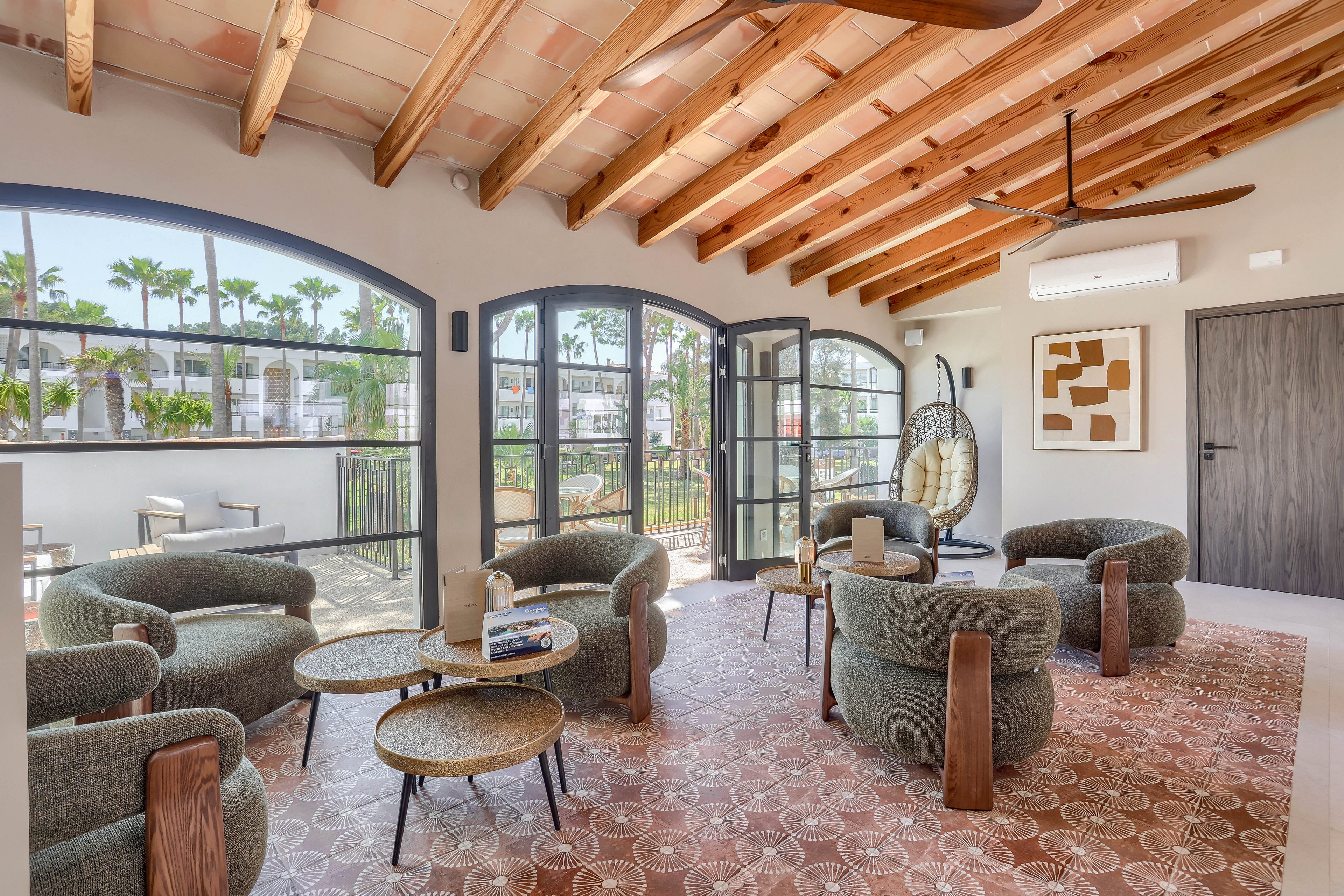 A stylish lounge area features curved armchairs, round coffee tables, and a hanging egg chair. The space is illuminated by large arched windows, revealing a lush outdoor garden with palm trees. The wooden beam ceiling and patterned tile floor add a warm, rustic touch, while contemporary decor and neutral tones create a relaxed atmosphere. Visible elements include books, cups, and abstract wall art.