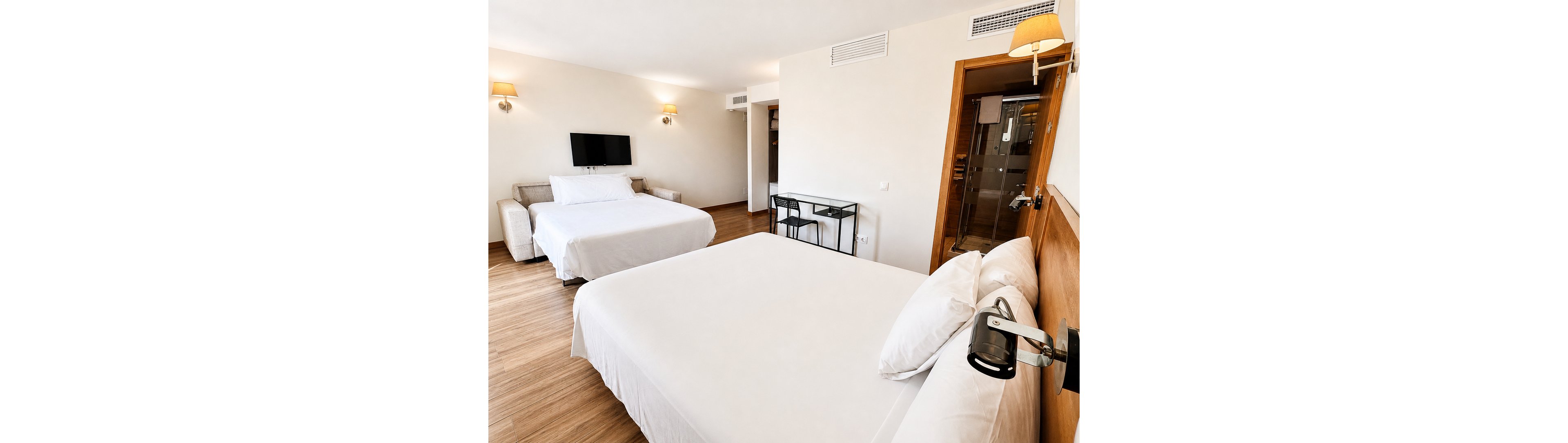 A spacious hotel room features two large neatly made beds with white linens. The interior has light-colored walls, wooden flooring, and minimal modern furniture including a small desk and chair. A flat-screen TV is mounted on the wall opposite one of the beds, and warm wall sconces provide soft lighting. A glass door reveals an adjoining bathroom area, enhancing the open and contemporary feel of the space.