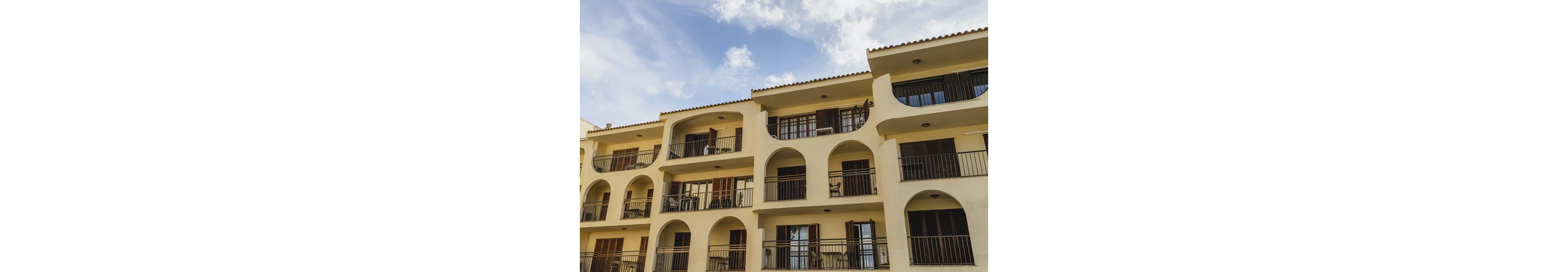 The image shows a multi-story apartment building featuring prominent arched balconies and railings. The structure is painted in a light beige color, with wooden doors and windows visible on each floor. The setting is outdoors under a partly cloudy sky, giving the scene a bright and airy mood. No visible text or numbers are present in the image.