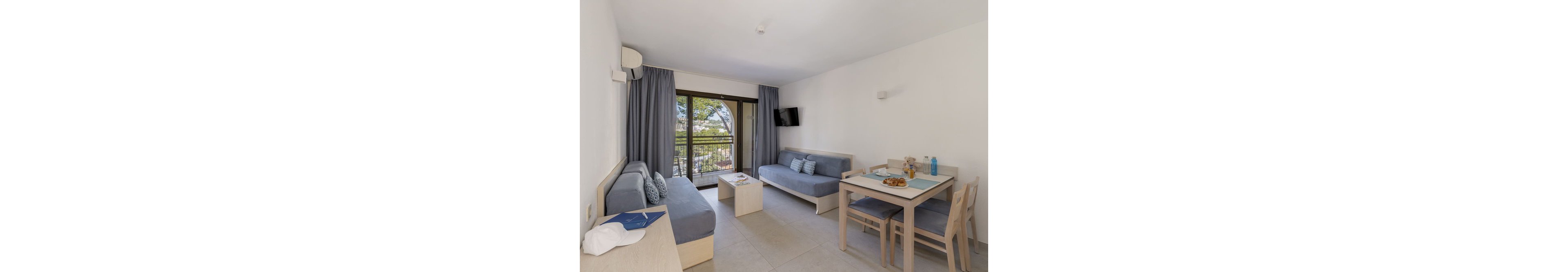 A contemporary apartment living room features two sofas, a dining table set for breakfast, and a wall-mounted television. The space is decorated in cool tones of blue and grey, with large windows and a balcony overlooking greenery. Bottled water and pastries are visible on the table, adding a welcoming touch to the bright, minimalist interior.