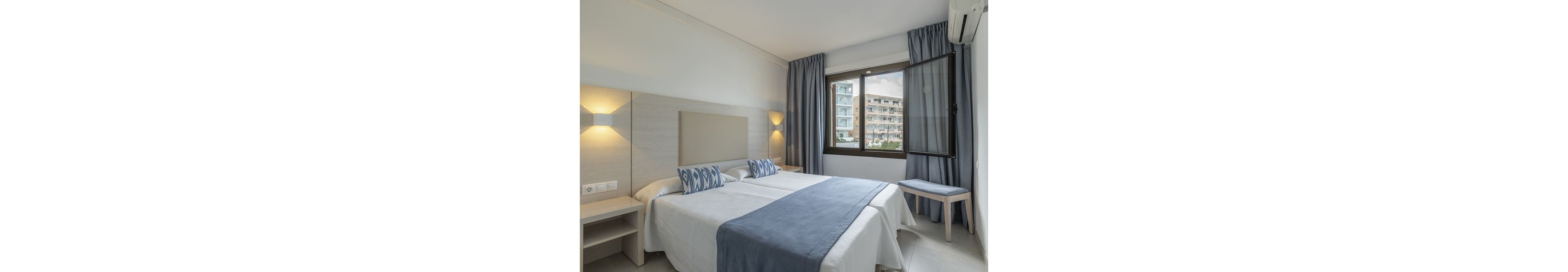 A contemporary hotel room features two twin beds with blue and white accent pillows and a matching bed runner. The space is bright, with neutral walls and soft lighting, and a large window offers a view of urban apartment buildings outside. Minimalist furniture, including a bedside table and bench, complements the clean and inviting atmosphere.