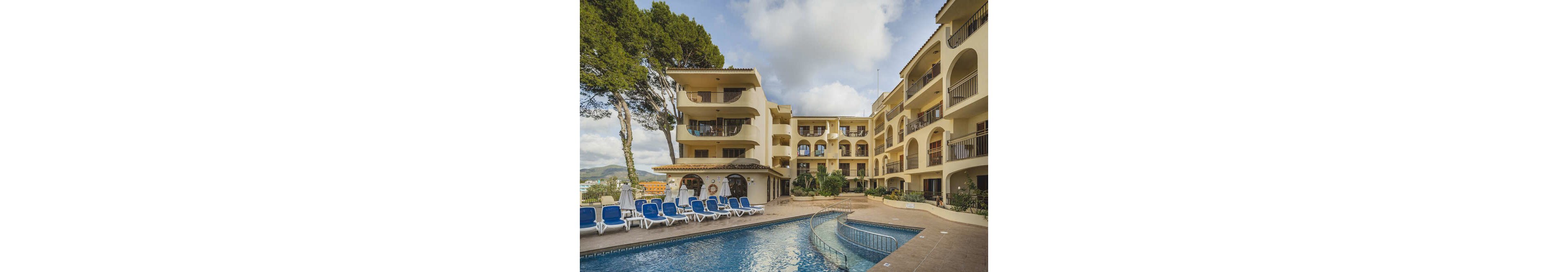 A multi-story Mediterranean style hotel features arched balconies and cream-colored walls. The outdoor area includes a swimming pool surrounded by blue and white lounge chairs, set against a backdrop of trees and partly cloudy skies. The tranquil setting suggests a relaxing vacation atmosphere, with no visible people or text.