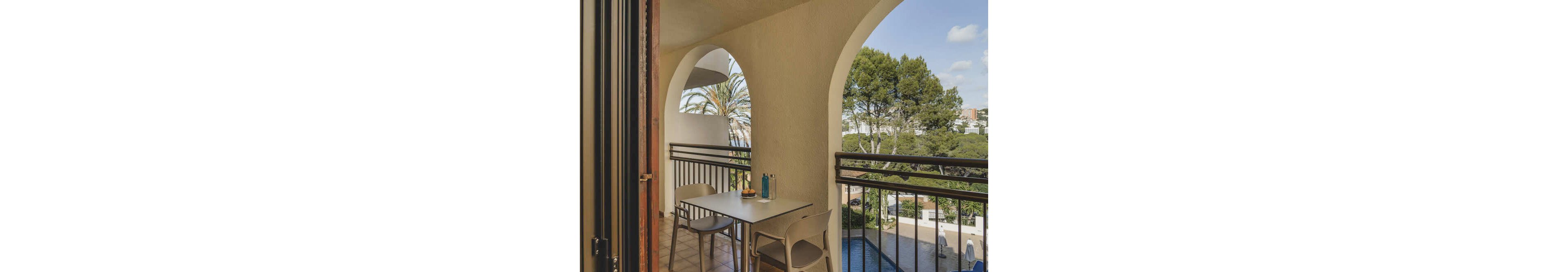 A small balcony features a modern table and two chairs, set against arched walls. The table holds two bottles and a plate, suggesting a casual meal or refreshment. The view includes lush green trees, a swimming pool below, and distant buildings, creating a relaxed outdoor atmosphere. Natural light and shadows enhance the tranquil mood.