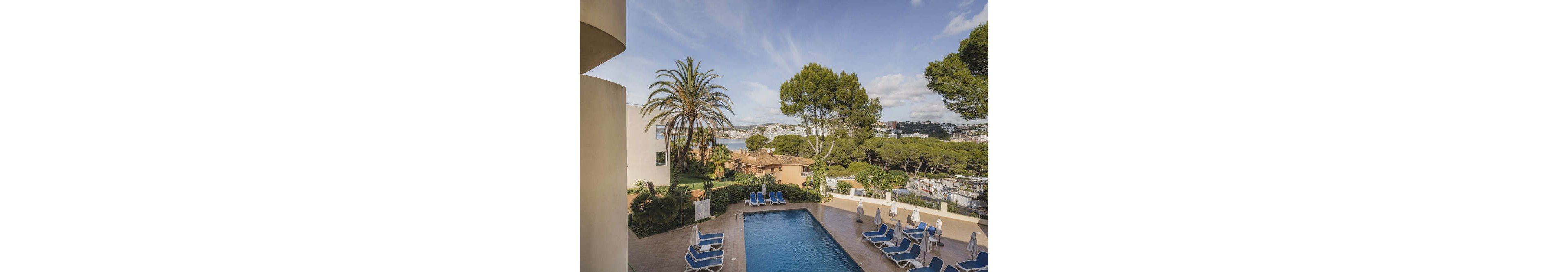 The main subject is a rectangular outdoor swimming pool surrounded by blue lounge chairs and umbrellas. The setting features lush greenery, palm trees, and a distant view of a coastal town with white buildings. The mood is relaxed and inviting, with a bright blue sky and soft natural lighting. No visible text or numeric values are present in the image.
