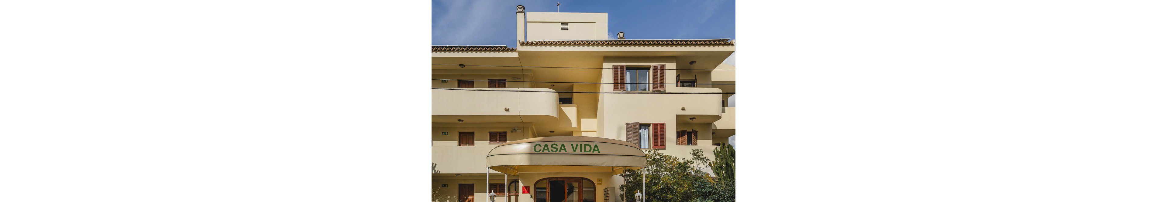The image shows the front facade of a building with the name 'CASA VIDA' clearly visible on an entrance awning. The architecture features balconies, shuttered windows, and a cream-colored exterior. The setting appears to be residential, with greenery and trees in the foreground. The lighting is natural, suggesting a sunny day. No people are visible in the scene.