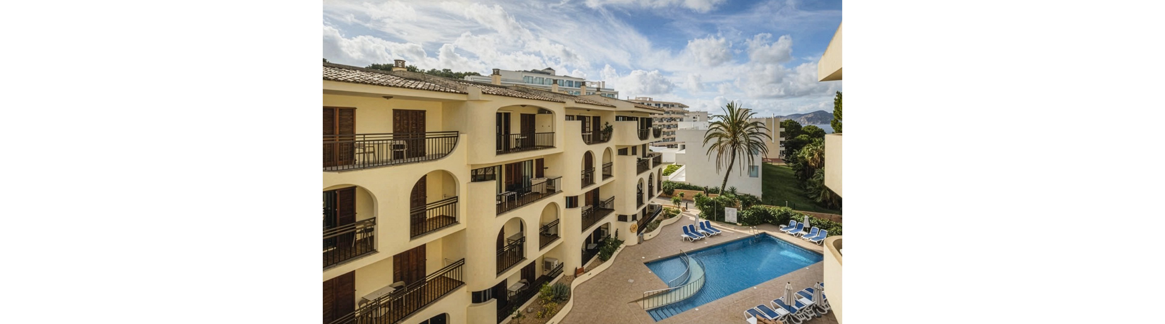 A multi-story Mediterranean-style hotel features arched balconies overlooking a central outdoor swimming pool. The setting is a sunny, resort-like environment with palm trees and lounge chairs arranged around the pool. The architecture is characterized by cream-colored walls and wooden shutters, creating a relaxed vacation atmosphere. The sky is partly cloudy, adding depth to the scene.