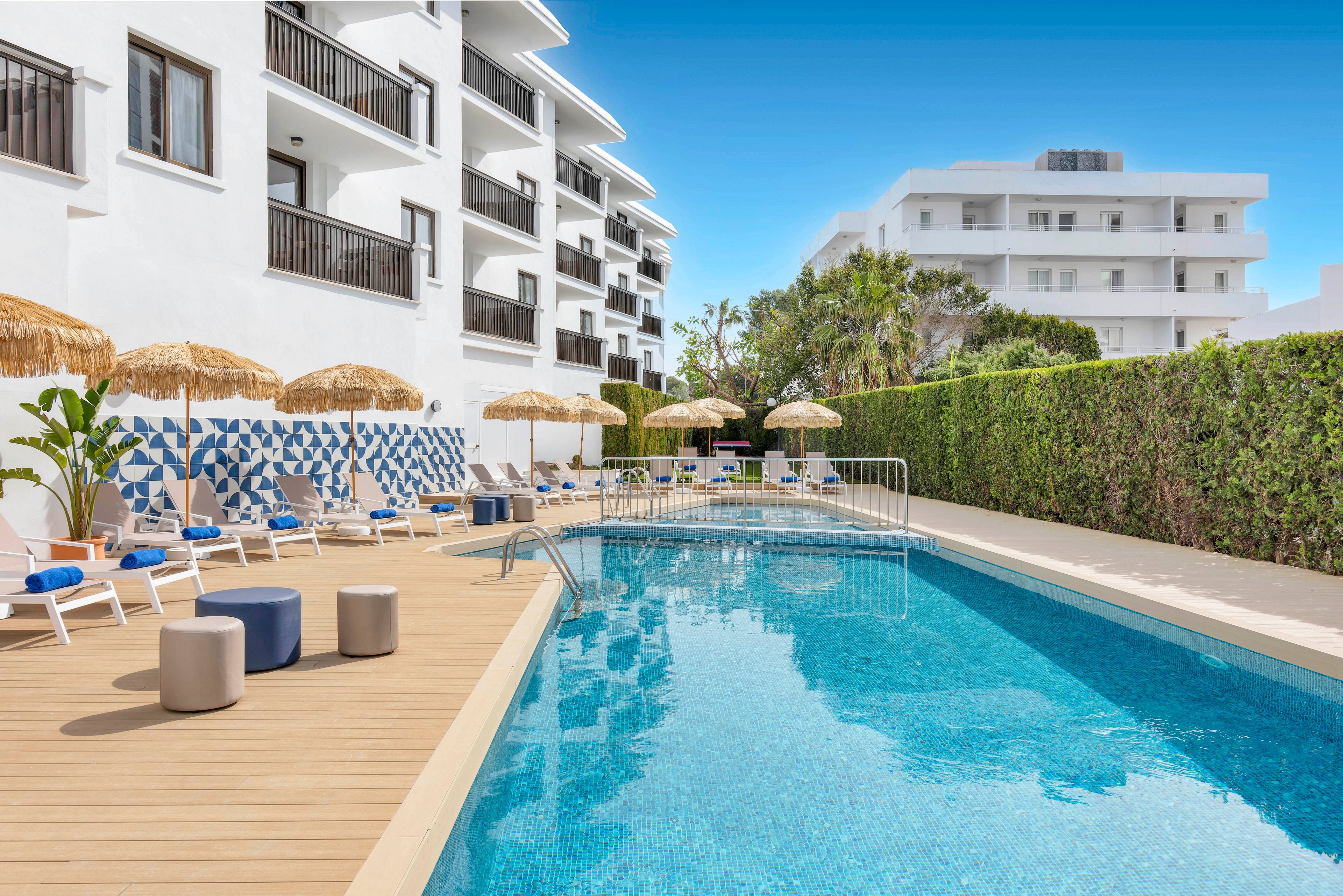 A clear blue swimming pool sits beside a modern white hotel building with balconies. Sun loungers and straw parasols line the wooden deck, creating a relaxed vacation atmosphere. Decorative blue-and-white wall tiles and potted plants add visual interest along the poolside. The scene is bright and sunny under a clear blue sky, suggesting a warm resort destination.