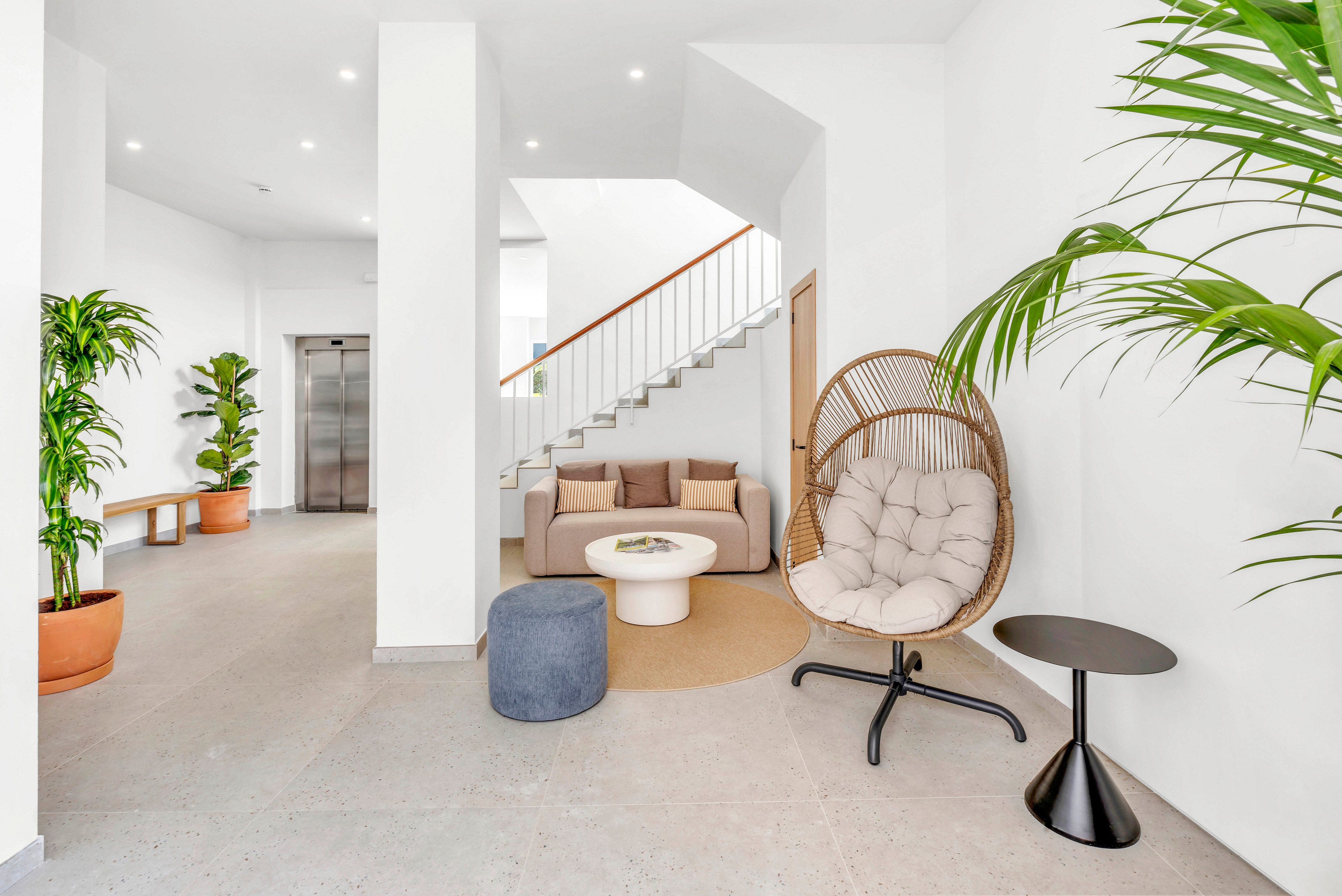A contemporary lobby features a neutral-toned sofa and round coffee table arranged on a light rug. A cushioned hanging chair and small black side table add stylish seating options near tall green potted plants. The space is bright and minimal, with white walls, recessed ceiling lights, and a staircase leading upward. An elevator and simple wooden bench are visible in the background, enhancing the clean, functional design.