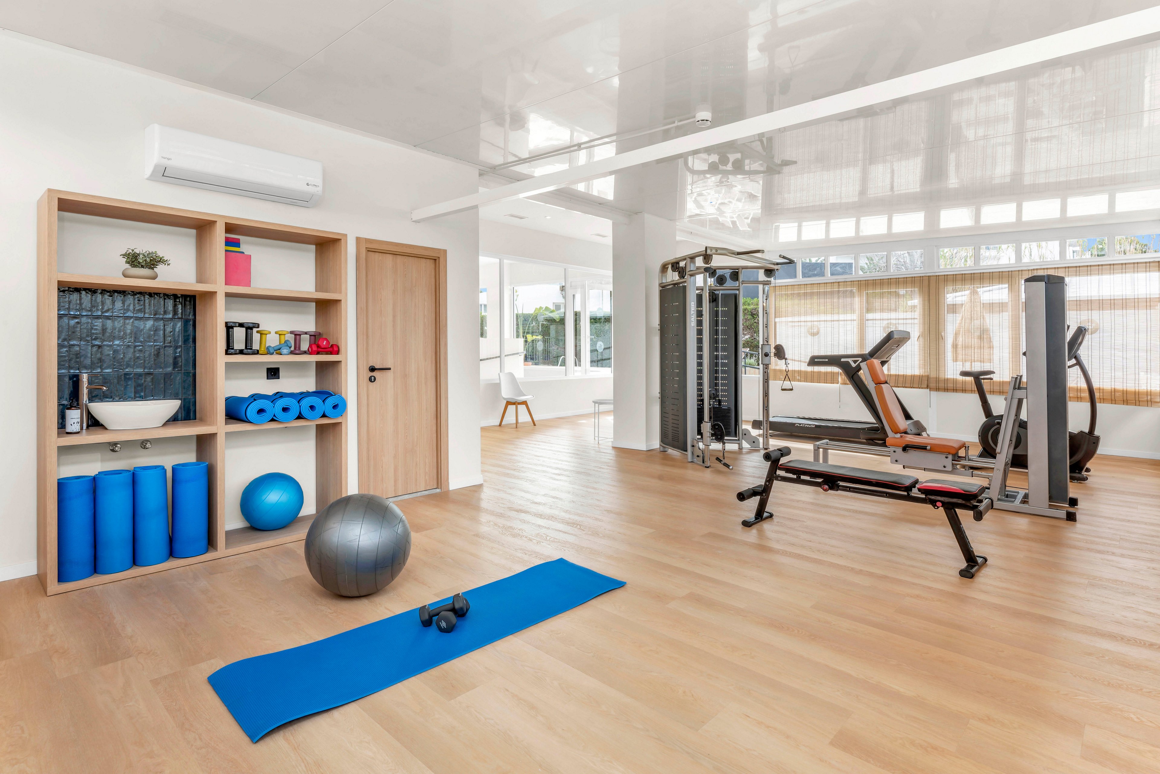 A spacious, modern home gym features various fitness equipment neatly arranged on a light wooden floor. In the foreground, a blue exercise mat, dumbbell, and silver stability ball create a workout area. Shelving on the left holds foam rollers, yoga mats, and small accessories, while strength machines and a weight bench stand on the right. Large windows and white walls give the room an airy, minimalist feel, ideal for personal training or wellness routines.