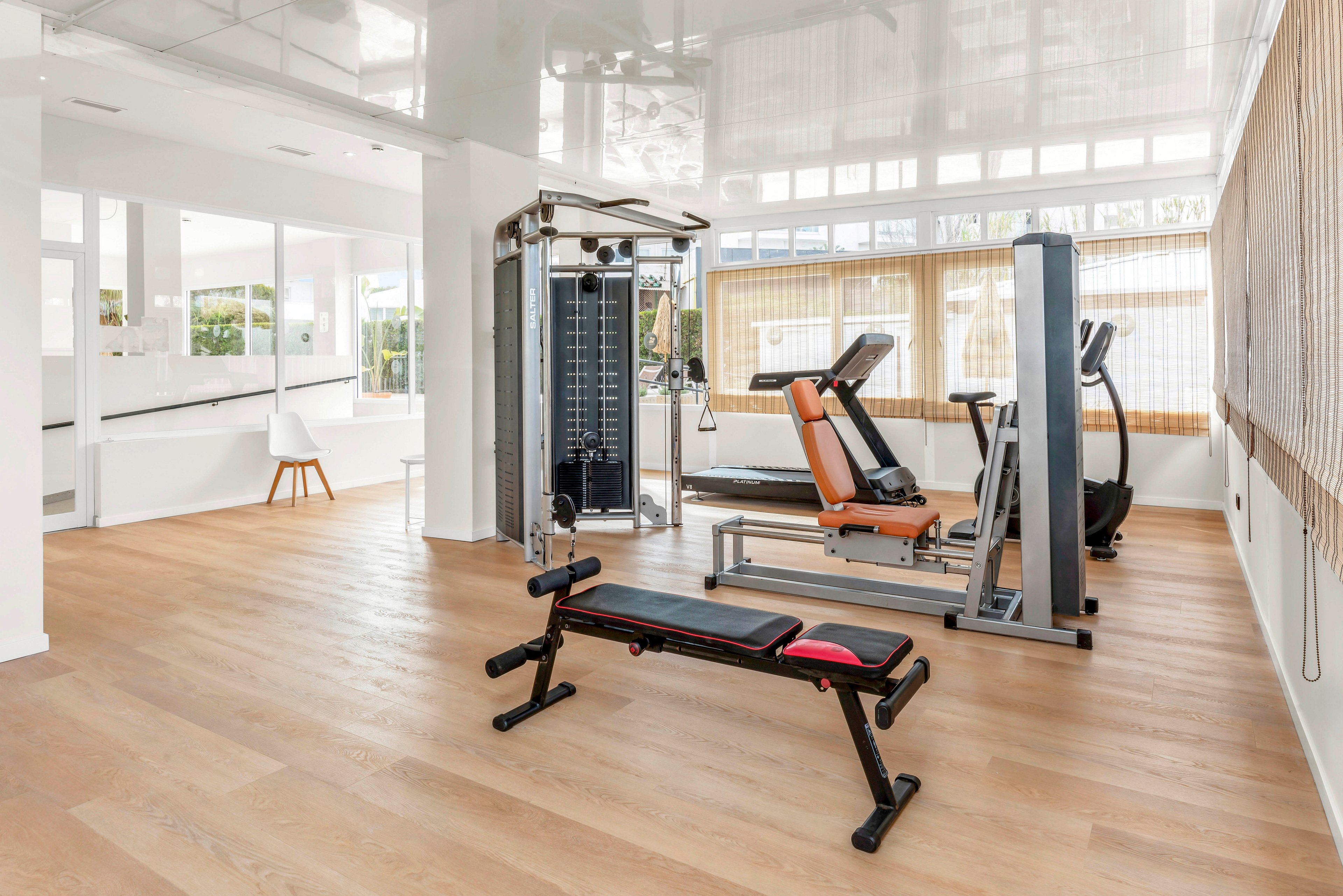 A clean, modern gym room features various fitness machines and a weight bench on a light wooden floor. Large windows and glass walls allow abundant natural light to fill the space, creating an open and airy atmosphere. The equipment includes a treadmill, strength training station, and padded bench arranged neatly for exercise. Neutral white walls and minimal decor emphasize a contemporary, uncluttered workout environment.