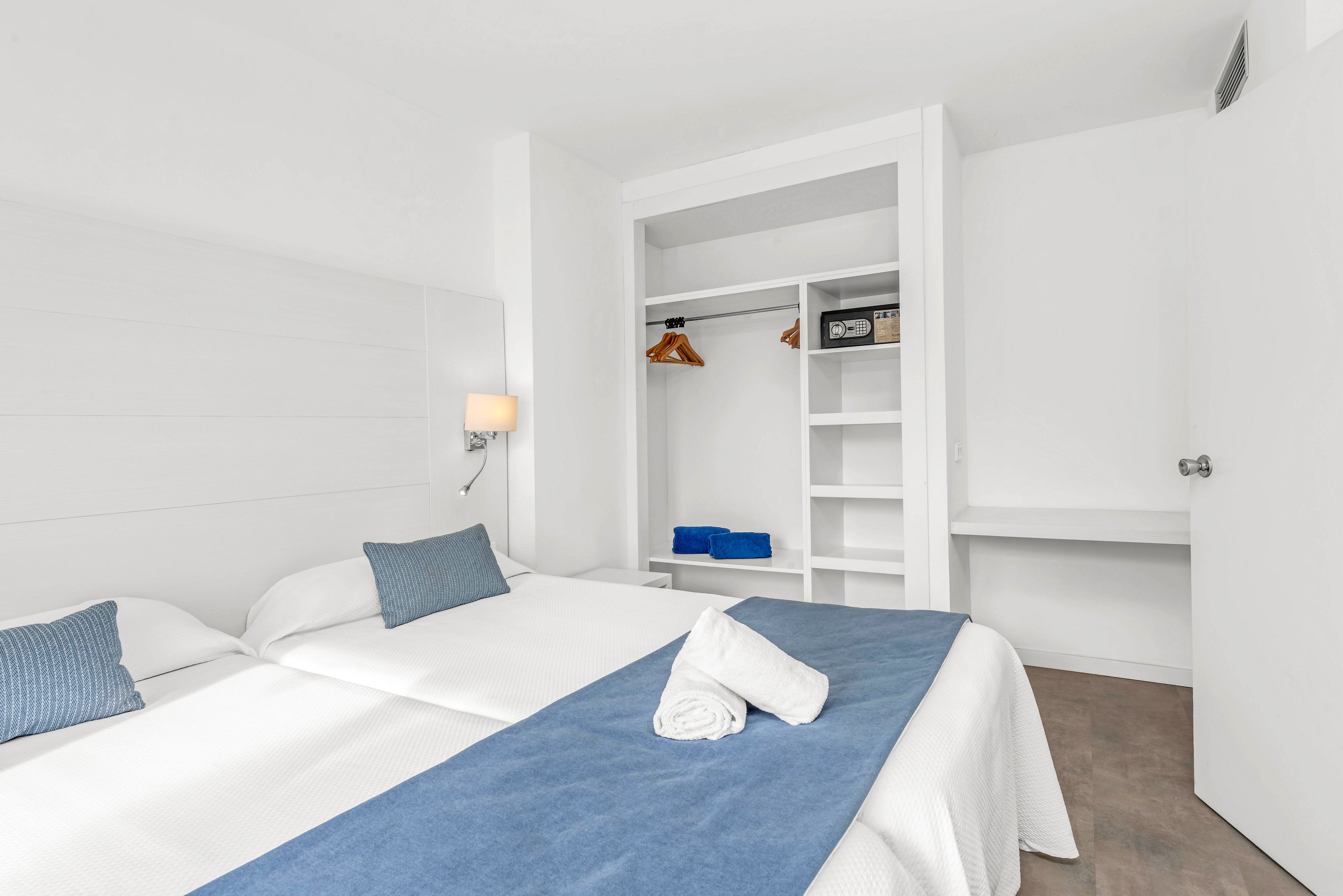 A bright white hotel room features two single beds pushed together with blue runners and matching pillows. An open built-in closet with shelves, hangers, and neatly folded blue towels is visible in the background. A wall-mounted bedside lamp adds soft lighting to the clean, minimalist space. The overall look is modern, tidy, and inviting.