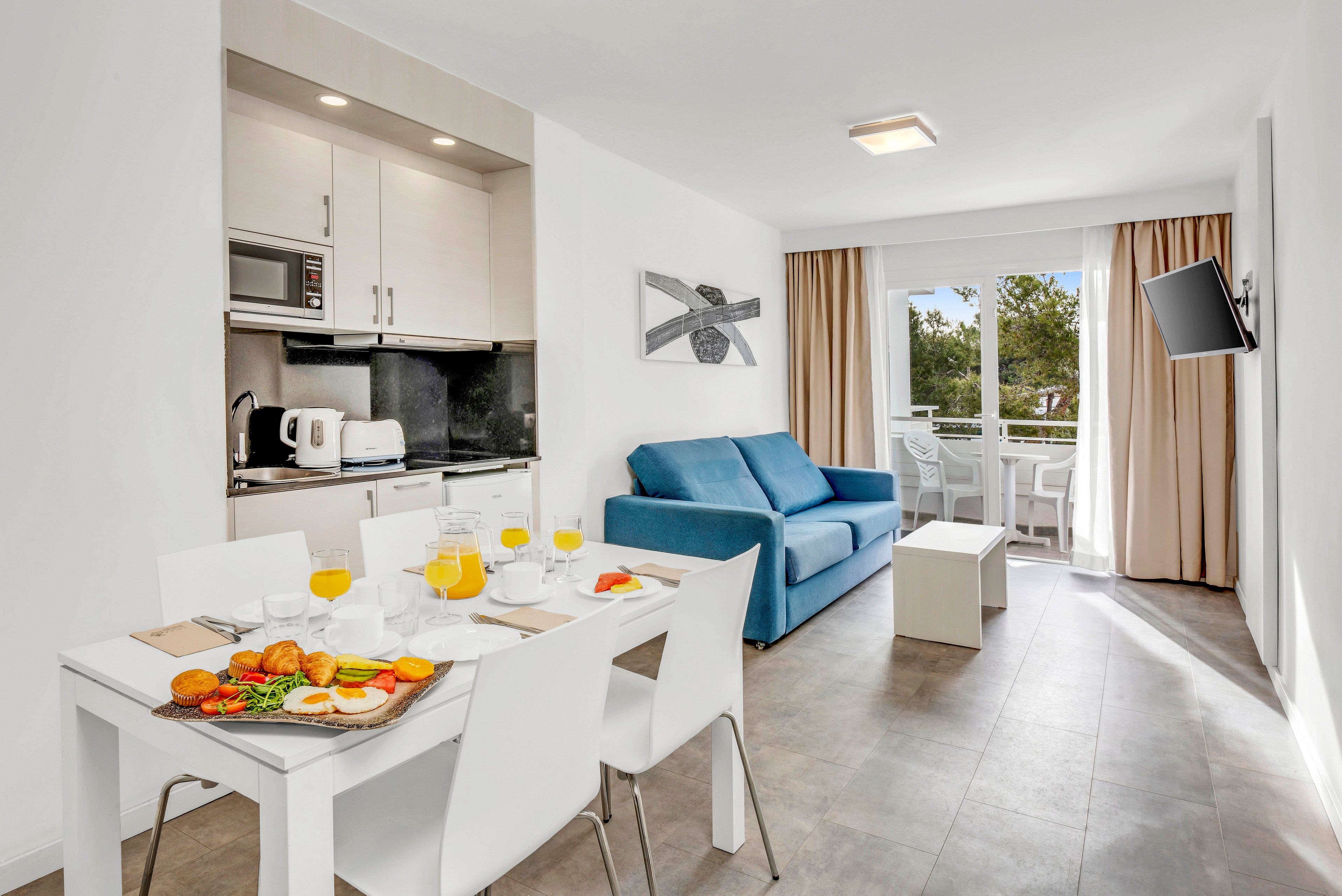 A contemporary open-plan apartment features a compact kitchen, dining area, and living room with a blue sofa. A white dining table is set with breakfast items, including croissants, fruit, and glasses of orange juice. The space is bright with white walls, minimalist furniture, and large windows leading to a balcony with outdoor seating. Neutral tones and clean lines create a fresh, inviting atmosphere suitable for vacation or short stays.