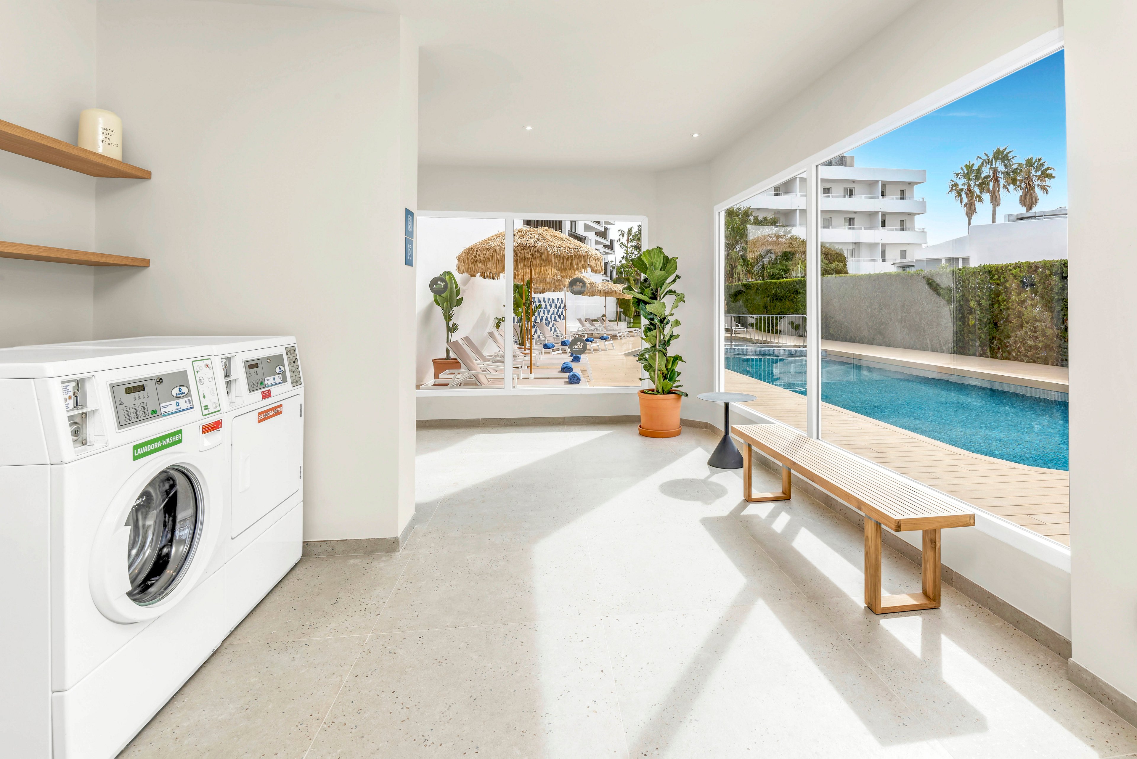 Bright contemporary laundry room features front-loading washing machines along one wall. Large floor-to-ceiling windows reveal a clear blue swimming pool and white apartment buildings outside. A wooden bench and potted plant add warmth to the minimalist interior. Sunlit patio umbrellas and palm trees in the background create a relaxed vacation atmosphere.