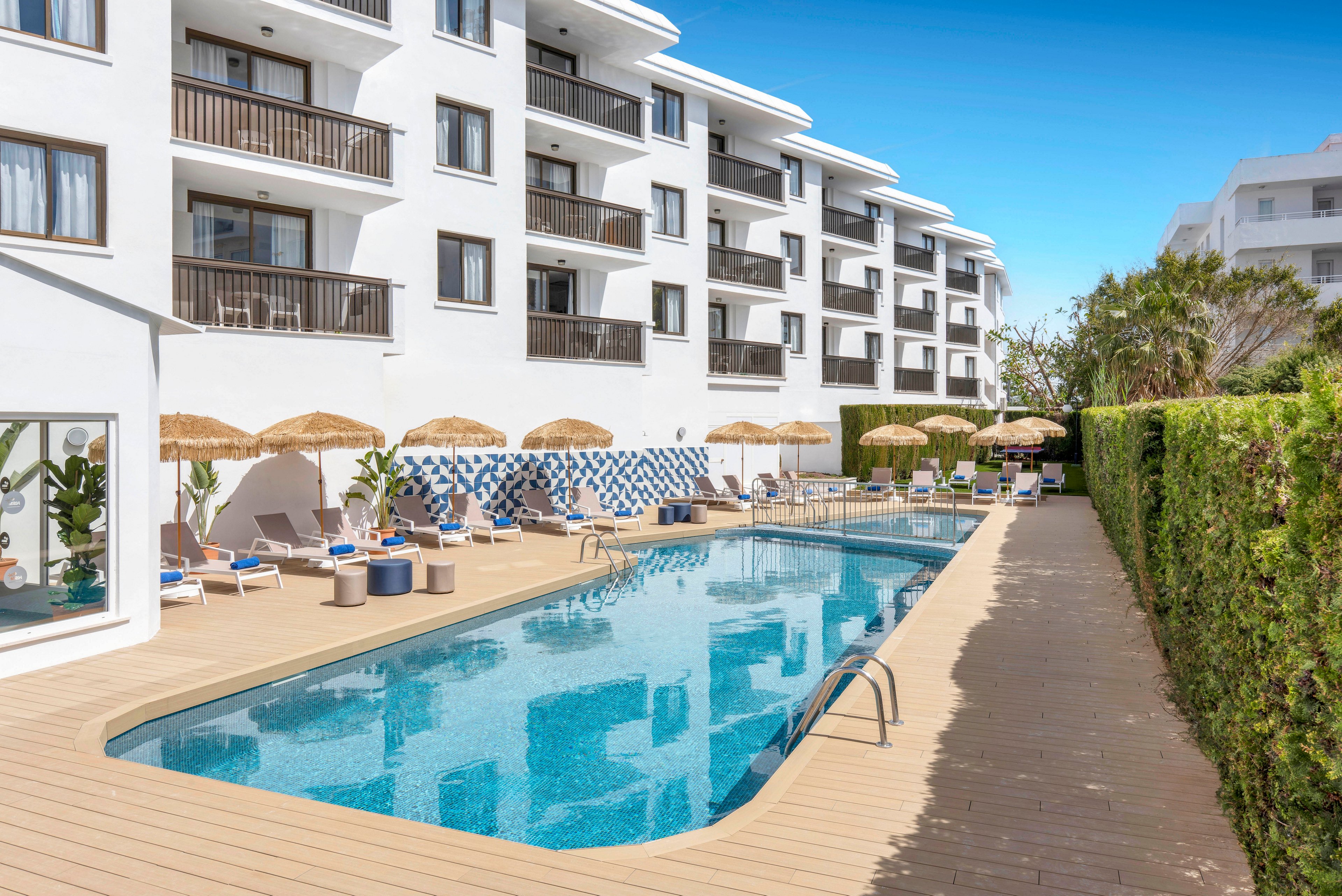 A contemporary white hotel building overlooks a long outdoor swimming pool. Sun loungers and straw parasols line the wooden deck, creating a relaxed vacation atmosphere. Blue and white tile accents, potted plants, and clear blue water add to the Mediterranean resort feel. A tall green hedge borders the pool area under a bright, cloudless sky.