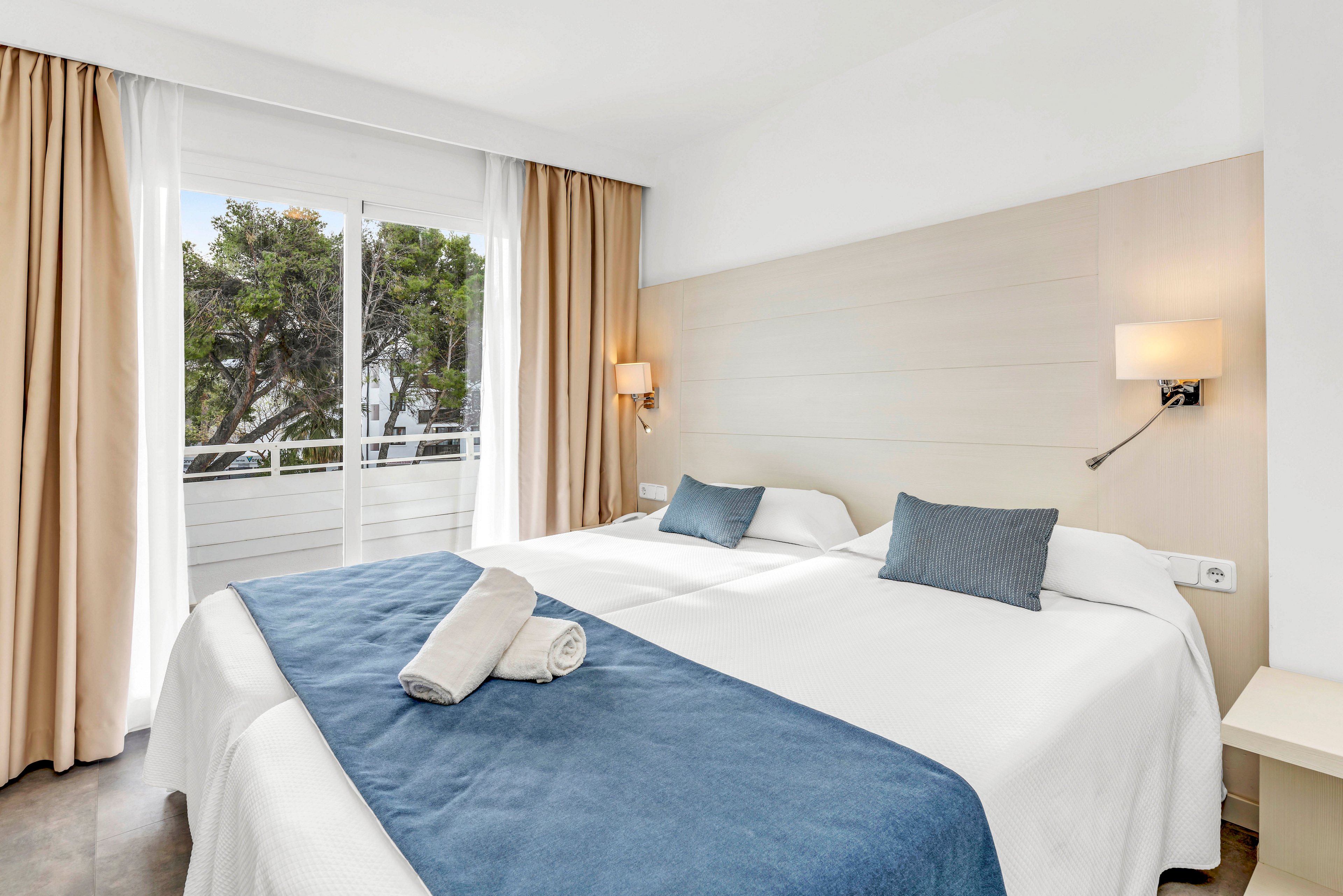 A clean, modern hotel room features two twin beds pushed together with white linens and blue accent runners. Two folded towels are neatly placed on the bed, suggesting readiness for guests. Natural light streams in through a large window with beige curtains, revealing trees and a balcony outside. Minimalist wall-mounted lamps and a small bedside table complete the calm, contemporary interior.