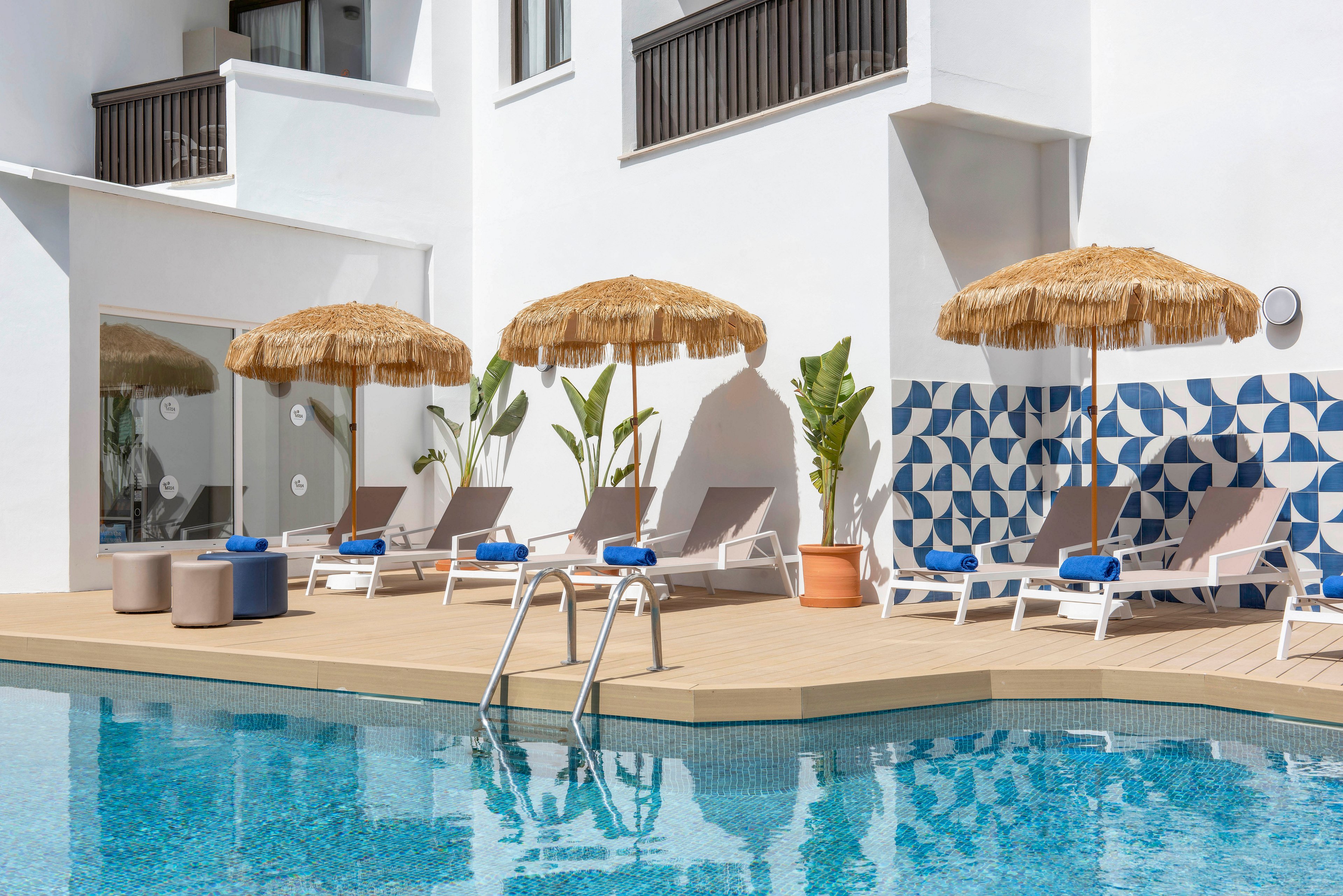 Contemporary outdoor swimming pool area at a white hotel building with several sun loungers arranged on a wooden deck. Straw parasols provide shade beside potted tropical plants and geometric blue-and-white wall tiles. Cushioned stools and rolled blue towels add comfort and color accents around the pool. The clear blue water reflects the clean architectural lines, creating a relaxed vacation atmosphere.