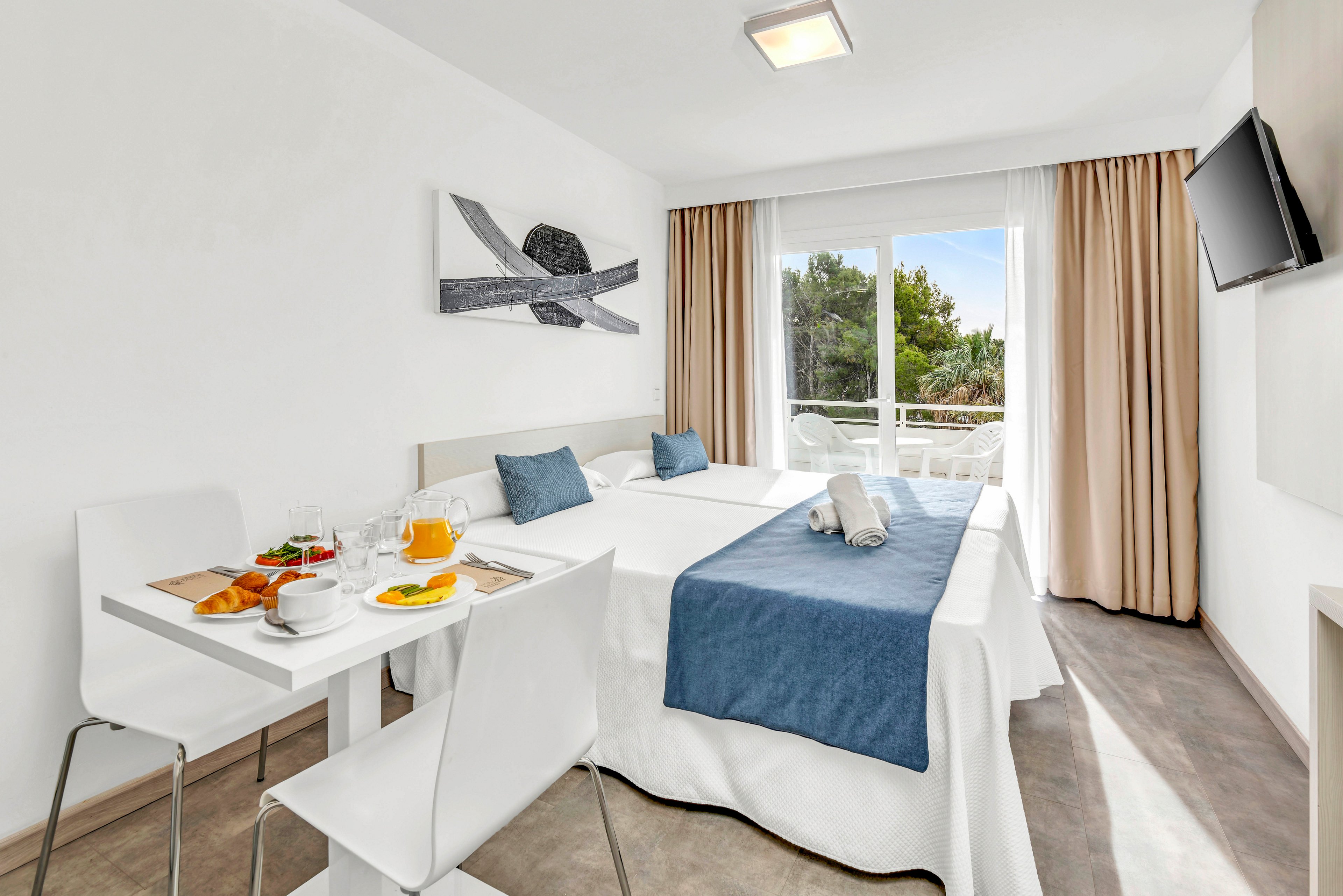 A bright, modern hotel room features a neatly made double bed with white linens and a blue runner. A small dining table is set with breakfast items including orange juice, coffee, and pastries. Large windows with beige curtains open to a balcony view of green trees, filling the space with natural light. Minimalist decor, including a wall-mounted television and abstract artwork, enhances the clean and contemporary atmosphere.