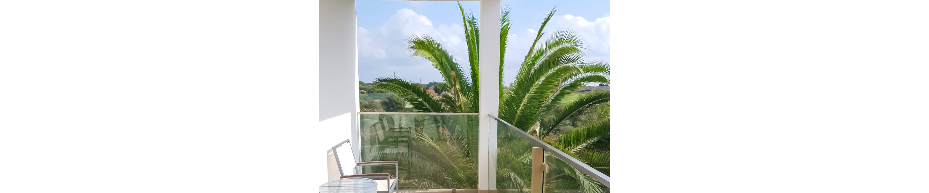 A serene balcony setting featuring a glass railing and a chair, overlooking lush palm trees and a scenic landscape. The sky is clear with soft clouds, creating a tranquil atmosphere. The setting suggests relaxation and outdoor leisure.