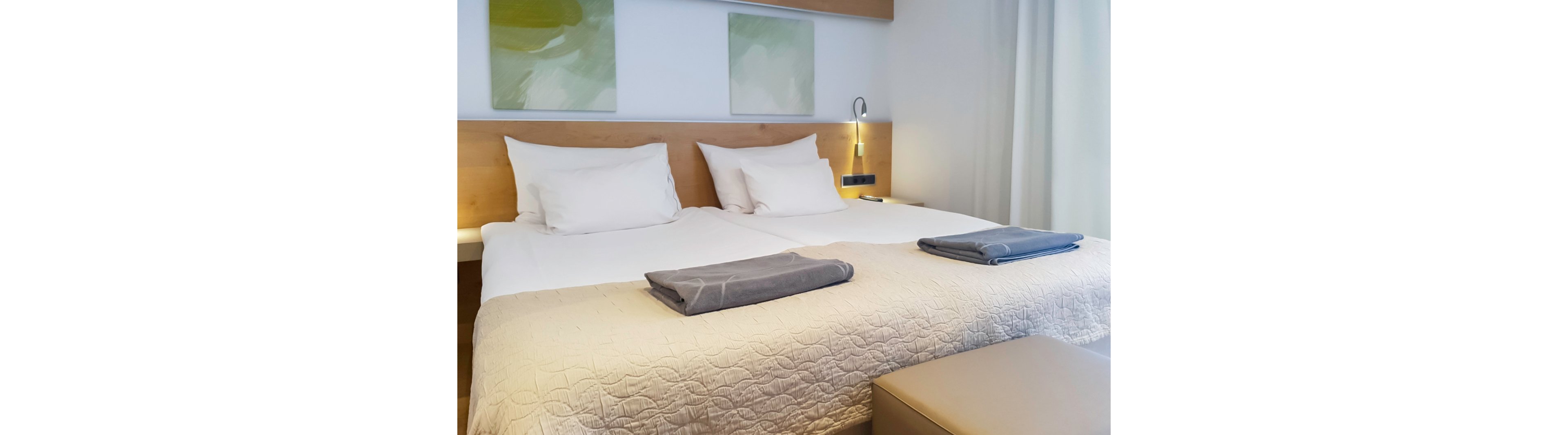 A contemporary hotel room featuring two neatly arranged twin beds with white linens and gray towels. The setting includes a wooden headboard, soft lighting, and abstract artwork on the wall. The room exudes a clean and minimalist aesthetic, ideal for relaxation or business stays.