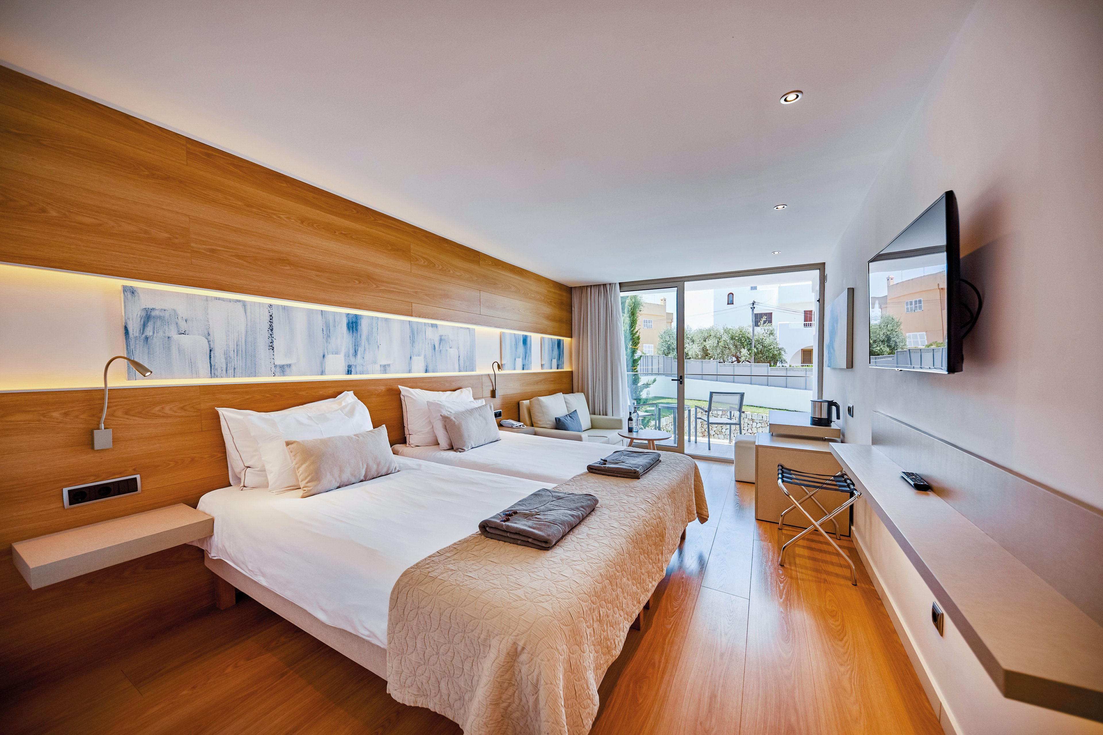 A contemporary hotel room featuring two neatly made beds with white linens and beige throws. The setting includes wooden paneling, abstract wall art, and a flat-screen TV mounted on the wall. A sliding glass door opens to a balcony with outdoor seating, overlooking a residential area. The room is well-lit with natural light and has a minimalist, cozy design.