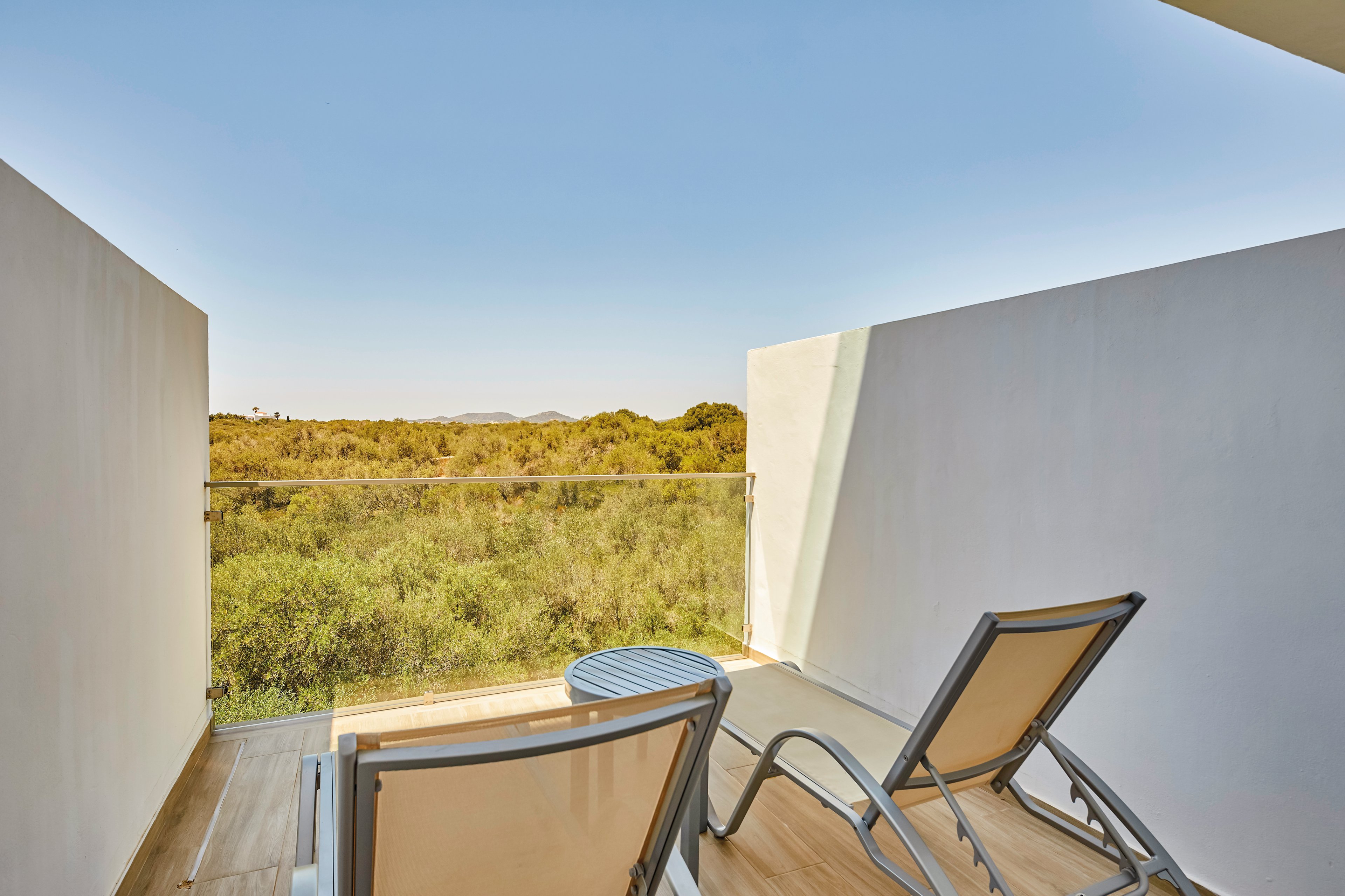 A serene balcony setting featuring two reclining lounge chairs facing a lush green landscape. The area is enclosed with a glass railing, offering an unobstructed view of nature. The scene is bathed in natural daylight under a clear blue sky, creating a tranquil and relaxing atmosphere.