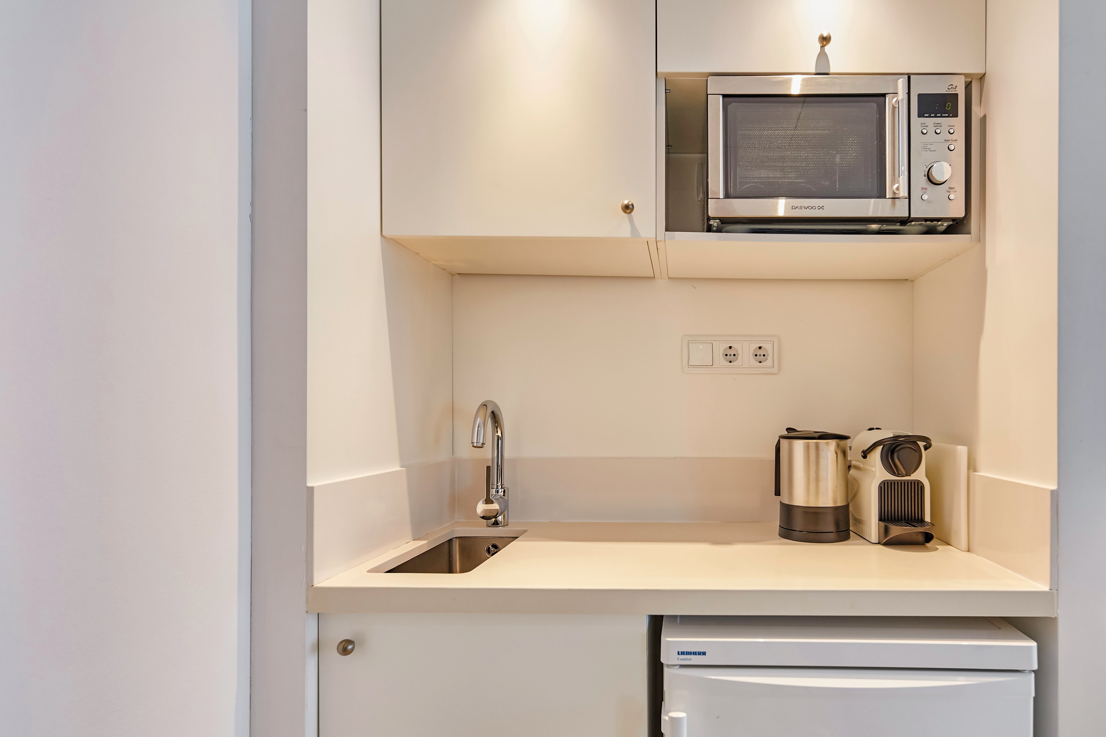 A small, modern kitchen setup featuring a microwave, electric kettle, and coffee machine. The space includes a stainless steel sink and a mini refrigerator, all set within a clean, white cabinetry design. The area is well-lit with built-in lighting, creating a functional and minimalist aesthetic.