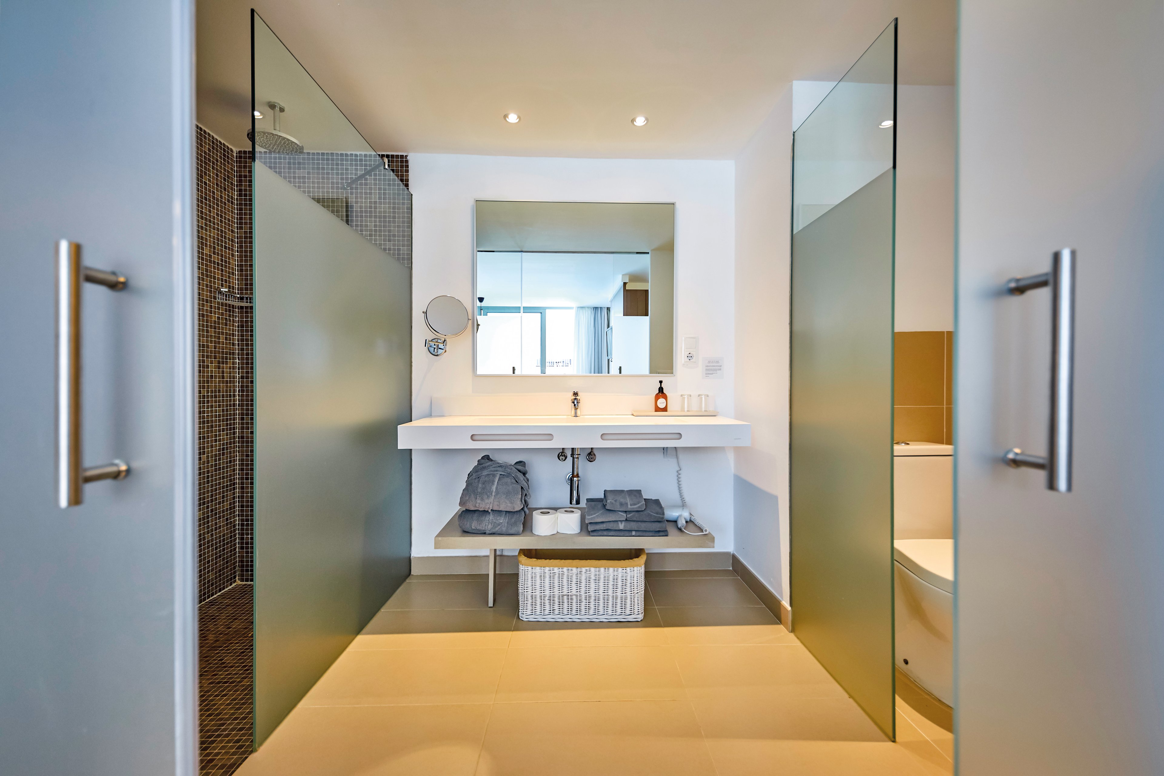 A contemporary bathroom featuring frosted glass partitions and a sleek white vanity. The setting includes neatly arranged towels and toiletries, with a wicker basket for storage. Bright lighting and a large mirror enhance the clean and minimalist design.