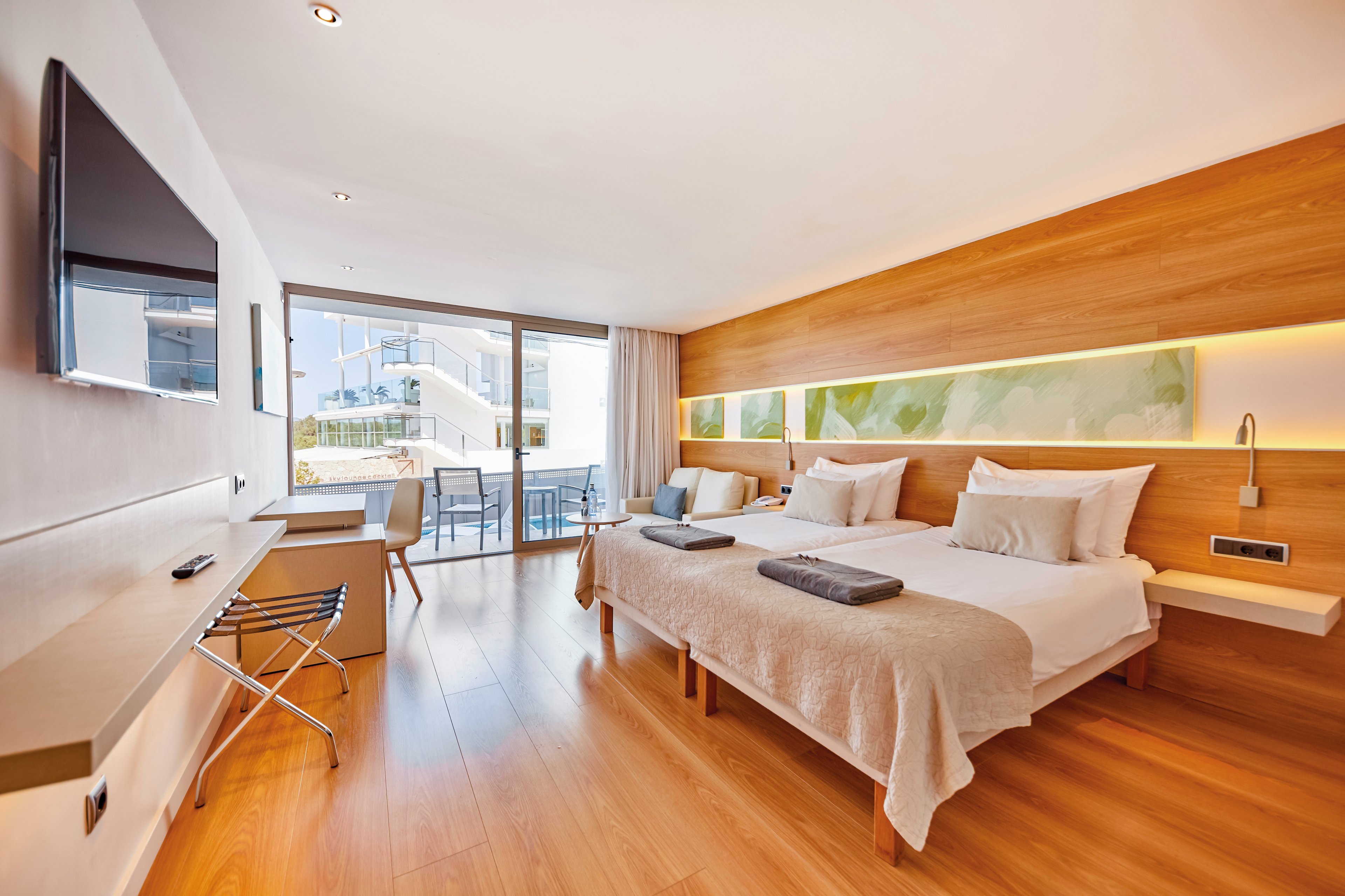 A spacious and contemporary hotel room featuring two neatly made beds with white linens and gray towels. The room includes wooden flooring, a wall-mounted TV, and a desk with a chair. Large glass doors open to a balcony overlooking a bright outdoor setting.