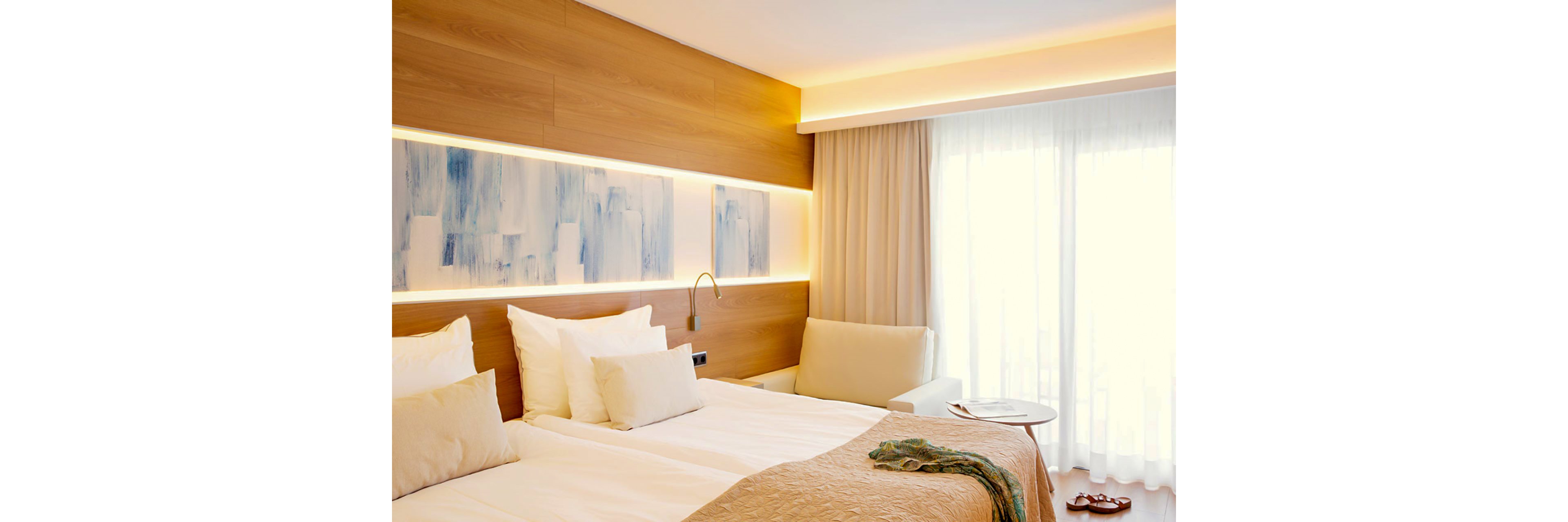 A bright and modern hotel room featuring a double bed with white linens and beige accents. The setting includes wooden paneling, abstract wall art, and soft ambient lighting. A seating area with a cream armchair and a small round table is visible near a large window with sheer curtains.