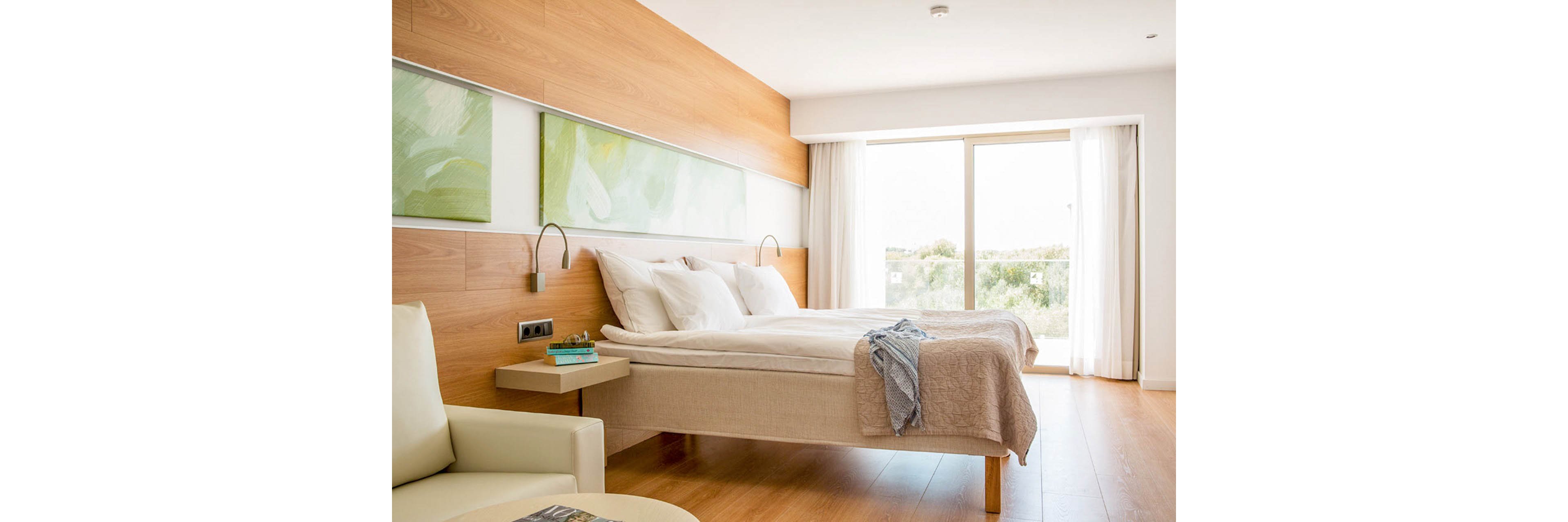 A bright, modern hotel room features two neatly made twin beds with white linens and a light throw draped over the foot. The space has warm wooden paneling, minimalist bedside tables, and built-in reading lights. Large windows with sheer curtains let in natural daylight and offer a view of greenery outside. Soft green artwork and neutral furniture create a calm, contemporary atmosphere.