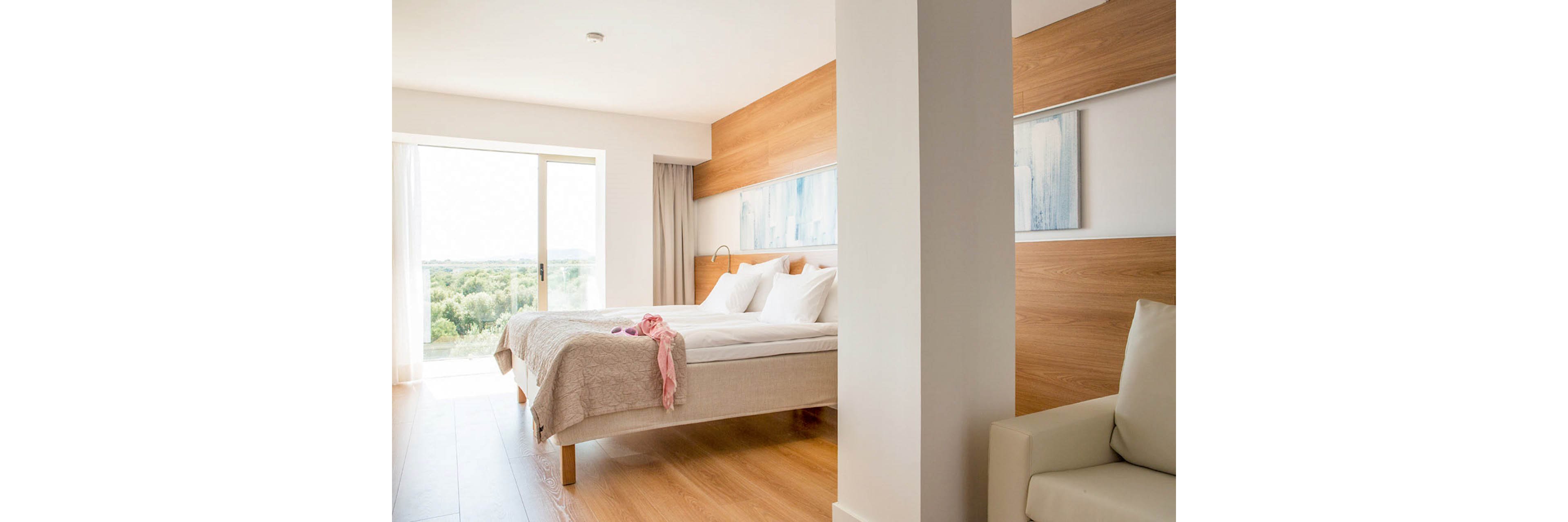 A bright, modern hotel bedroom features a neatly made double bed with white linens and a light beige bedspread. Soft wooden flooring and wood-paneled walls create a warm, minimalist aesthetic. A sliding glass door opens to a balcony with a scenic view of greenery and distant hills. Neutral tones, simple artwork, and natural light give the room a calm, inviting atmosphere.