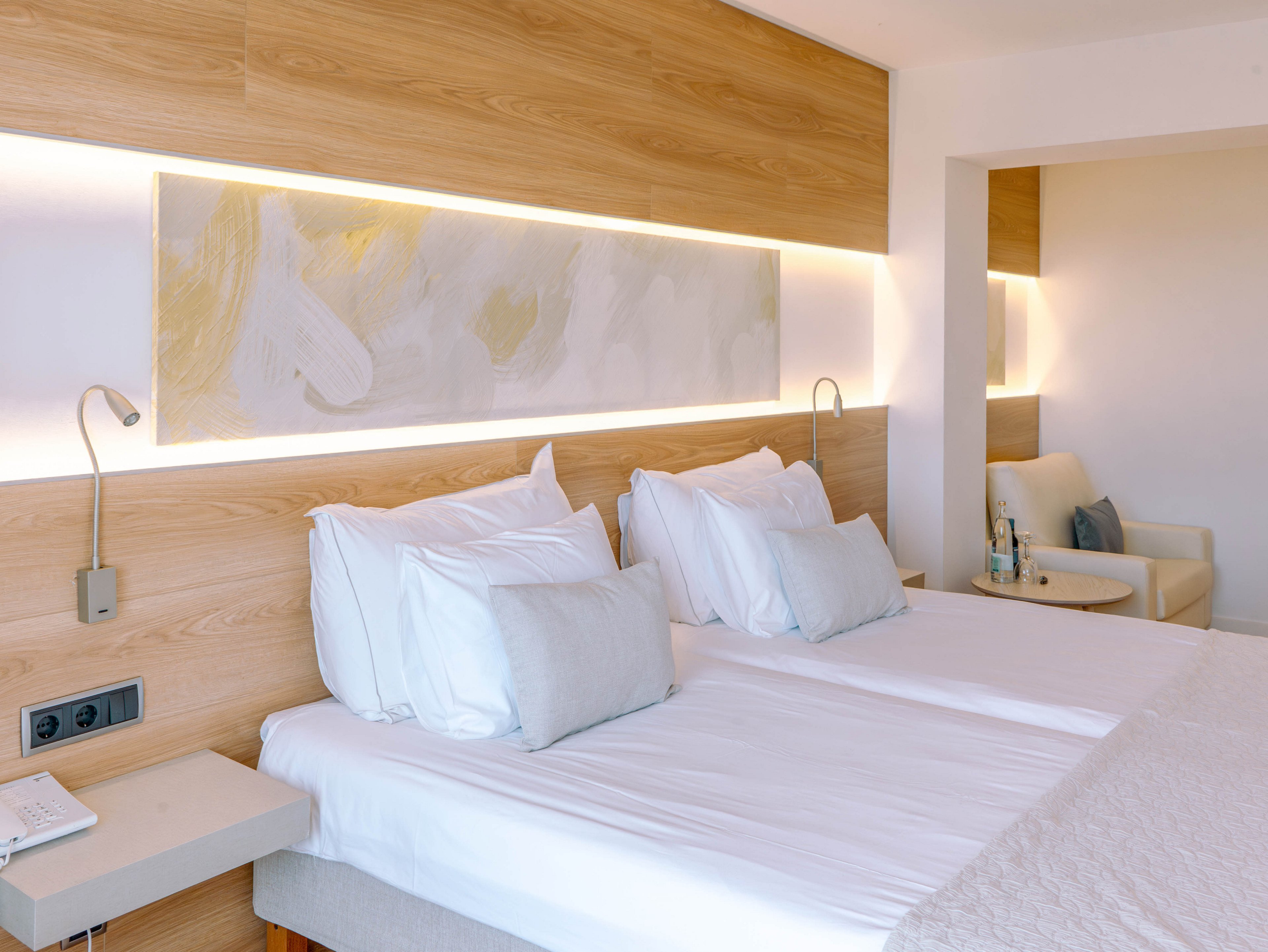 A bright modern hotel room features two neatly made twin beds with crisp white linens and light gray accent pillows. Warm wooden paneling and a long illuminated wall art piece create a cozy, contemporary atmosphere. Built-in reading lights and a small bedside table add functional comfort. In the background, a lounge chair and side table with bottled water complete the relaxing setting.