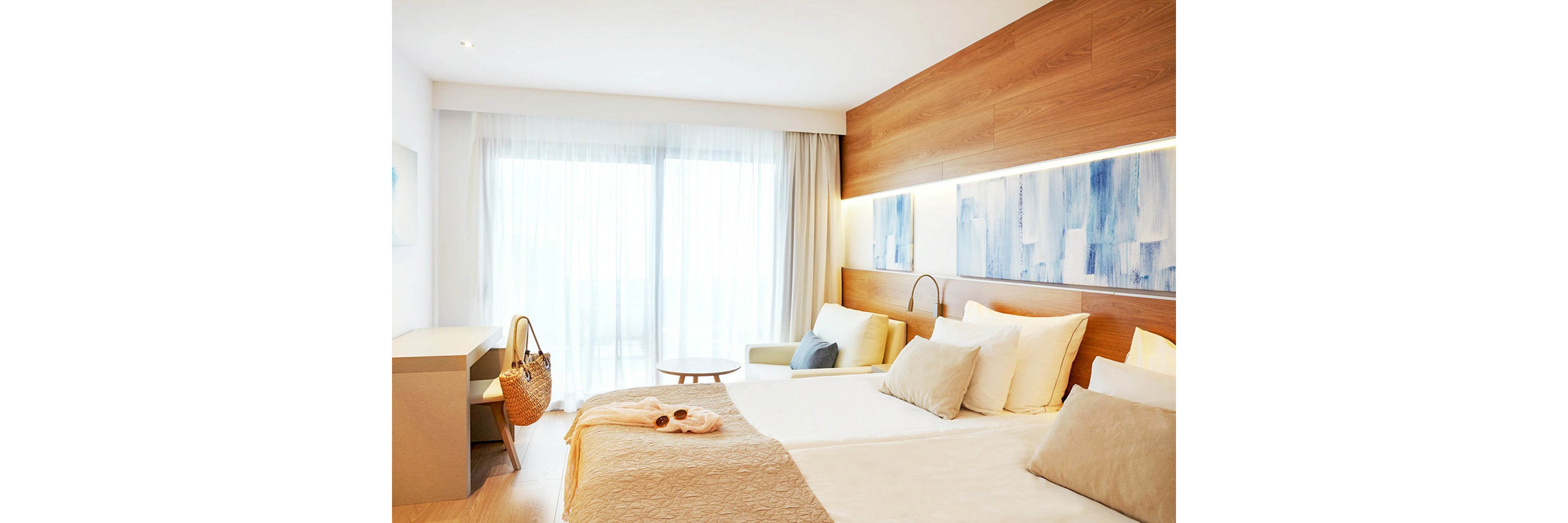 A bright, modern hotel bedroom features a neatly made double bed with soft neutral bedding and multiple pillows. Floor-to-ceiling sheer curtains cover a large glass balcony door, allowing diffused daylight to fill the room. A small sofa with cushions, a minimalist desk, and a round side table add comfort and functionality. Warm wooden wall panels and abstract blue artwork create a calm, contemporary atmosphere, while a woven bag on the bed suggests a relaxed vacation setting.