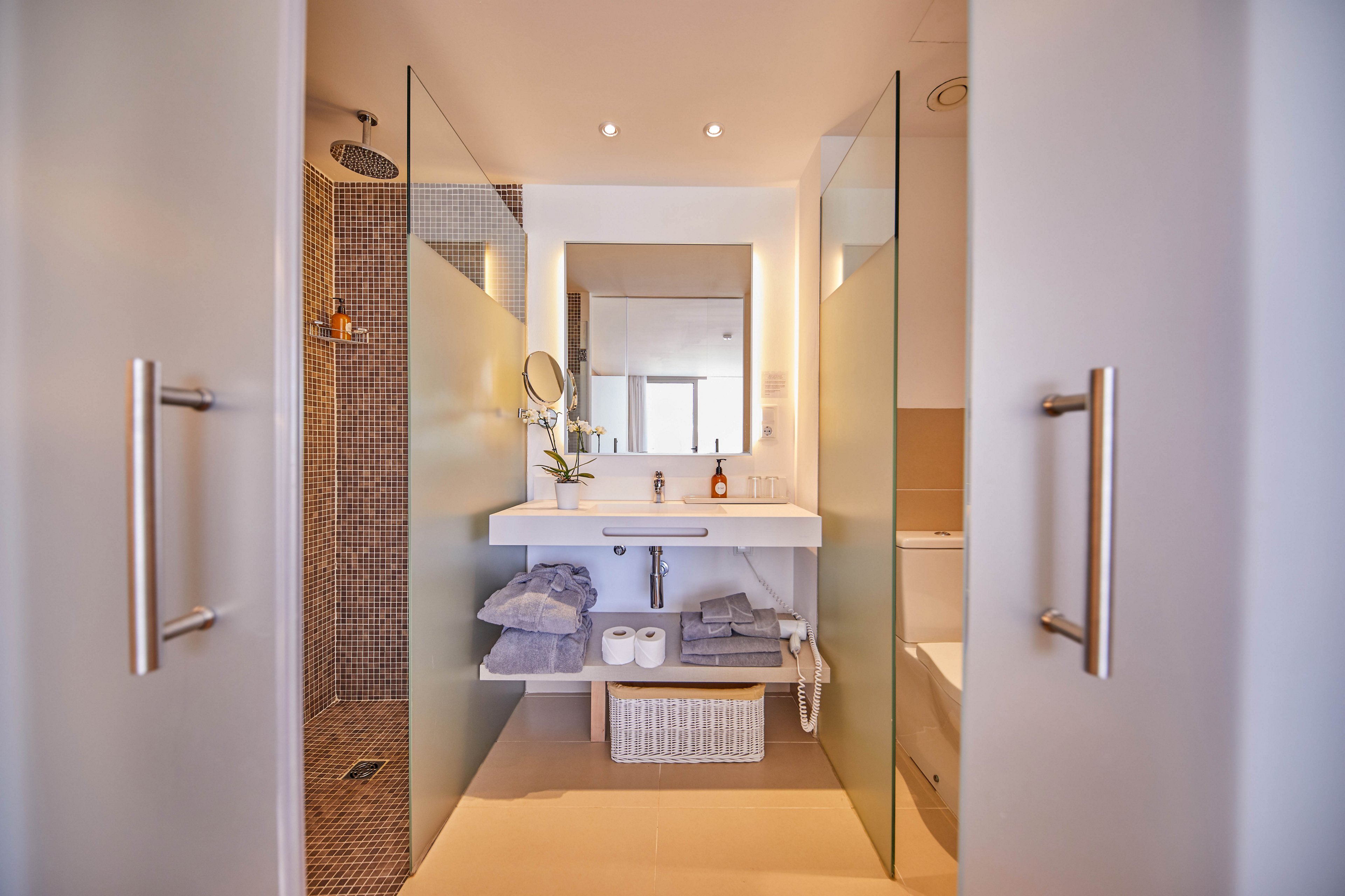 A contemporary bathroom features a sleek white vanity with an integrated sink and mirror. Frosted glass partitions frame the space, leading toward a walk-in shower with mosaic tiles. Soft towels, a wicker basket, and neatly arranged toiletries add a sense of order and comfort. Warm lighting and neutral tones create a calm, spa-like atmosphere.
