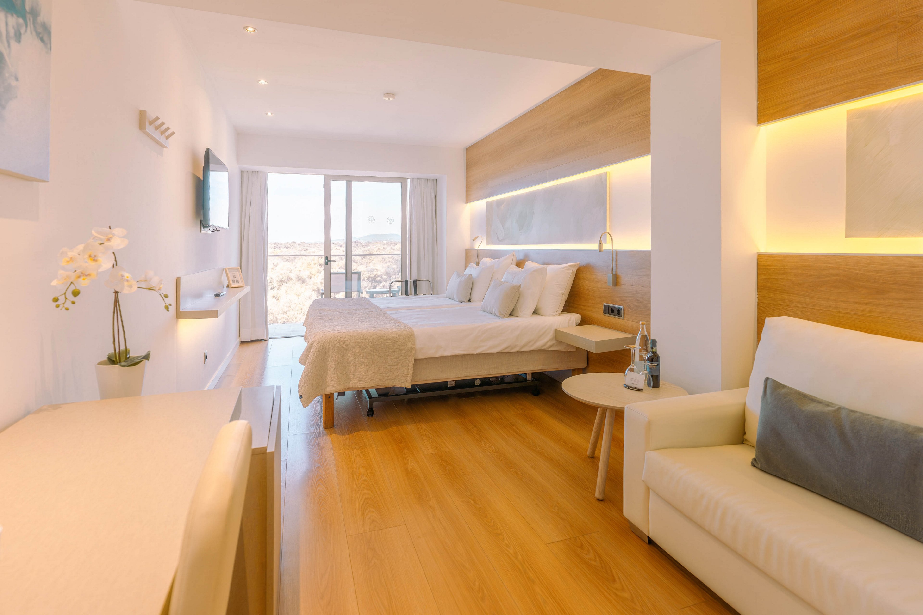 A spacious, contemporary hotel room features a large bed with white linens and soft pillows. Warm wooden flooring and wall panels contrast with white walls and furniture, including a sofa and desk. Recessed lighting and LED strip lights create a cozy, inviting atmosphere. Floor-to-ceiling glass doors open to a balcony with a distant city or coastal view, filling the room with natural light.
