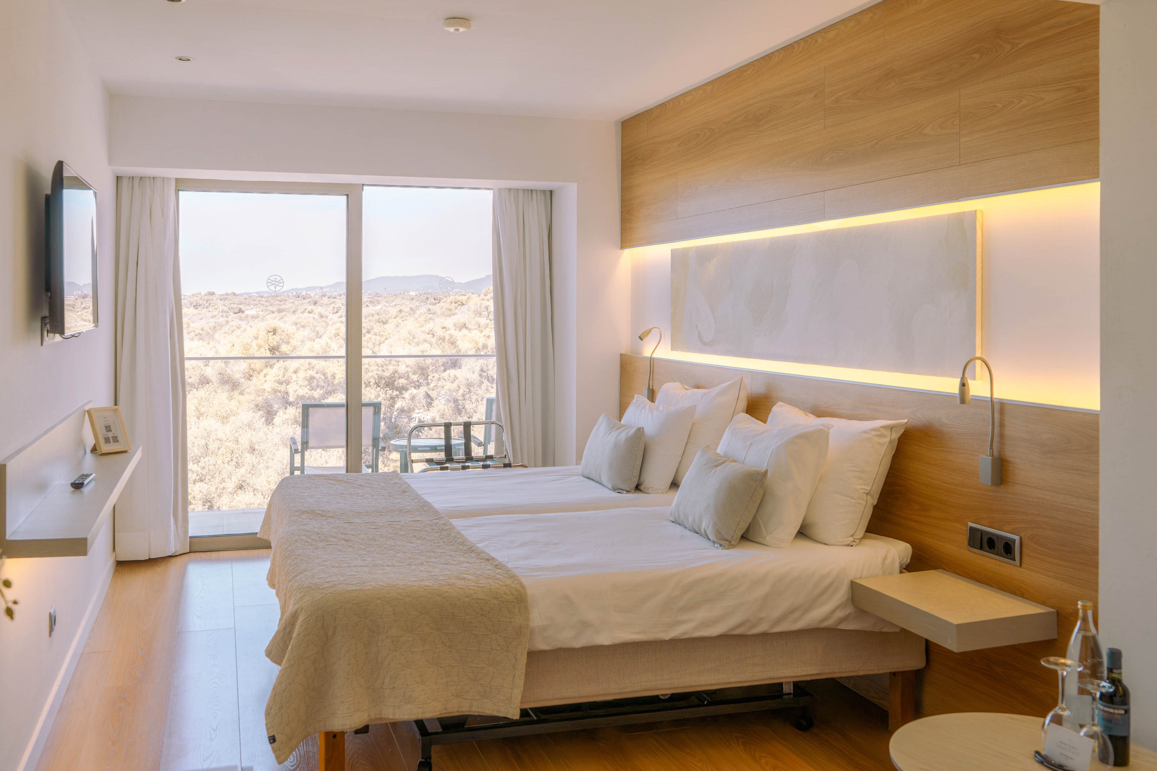 A contemporary hotel bedroom features two neatly made beds with white linens and light beige throws. Warm wooden paneling and integrated strip lighting create a cozy, minimalist atmosphere. A large glass door opens to a balcony overlooking a dry, desert-like landscape. Subtle furnishings, including wall-mounted lamps, a small side table, and a flat-screen TV, complete the clean, modern design.