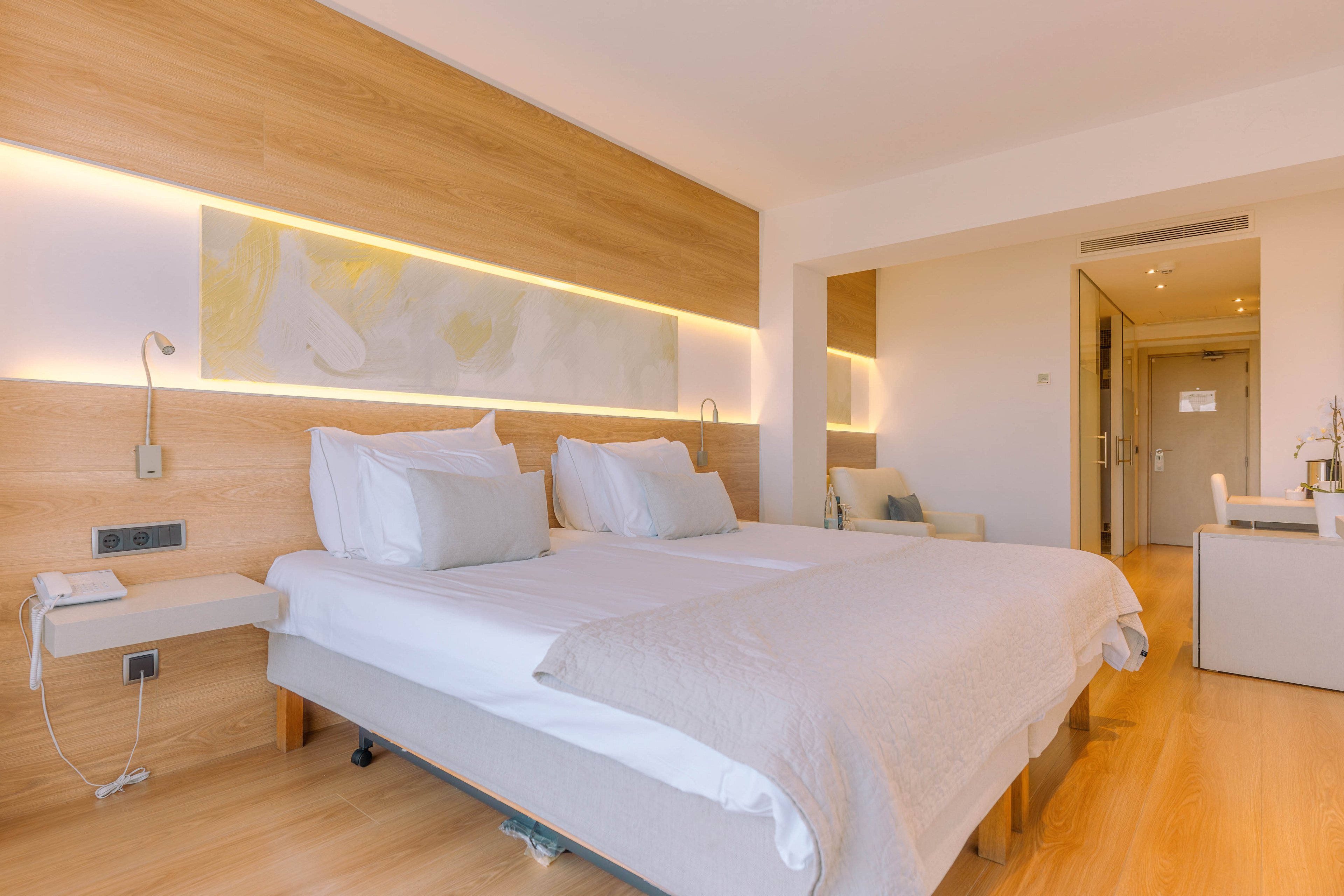 A bright, modern hotel room features a large double bed with crisp white linens and neutral-toned bedding. Warm wooden paneling with integrated strip lighting runs along the headboard wall, adding a soft ambient glow. A minimalist desk area and chair are visible near the far wall, along with a doorway leading to the hall. The overall design is clean, contemporary, and inviting, emphasizing comfort and simplicity.