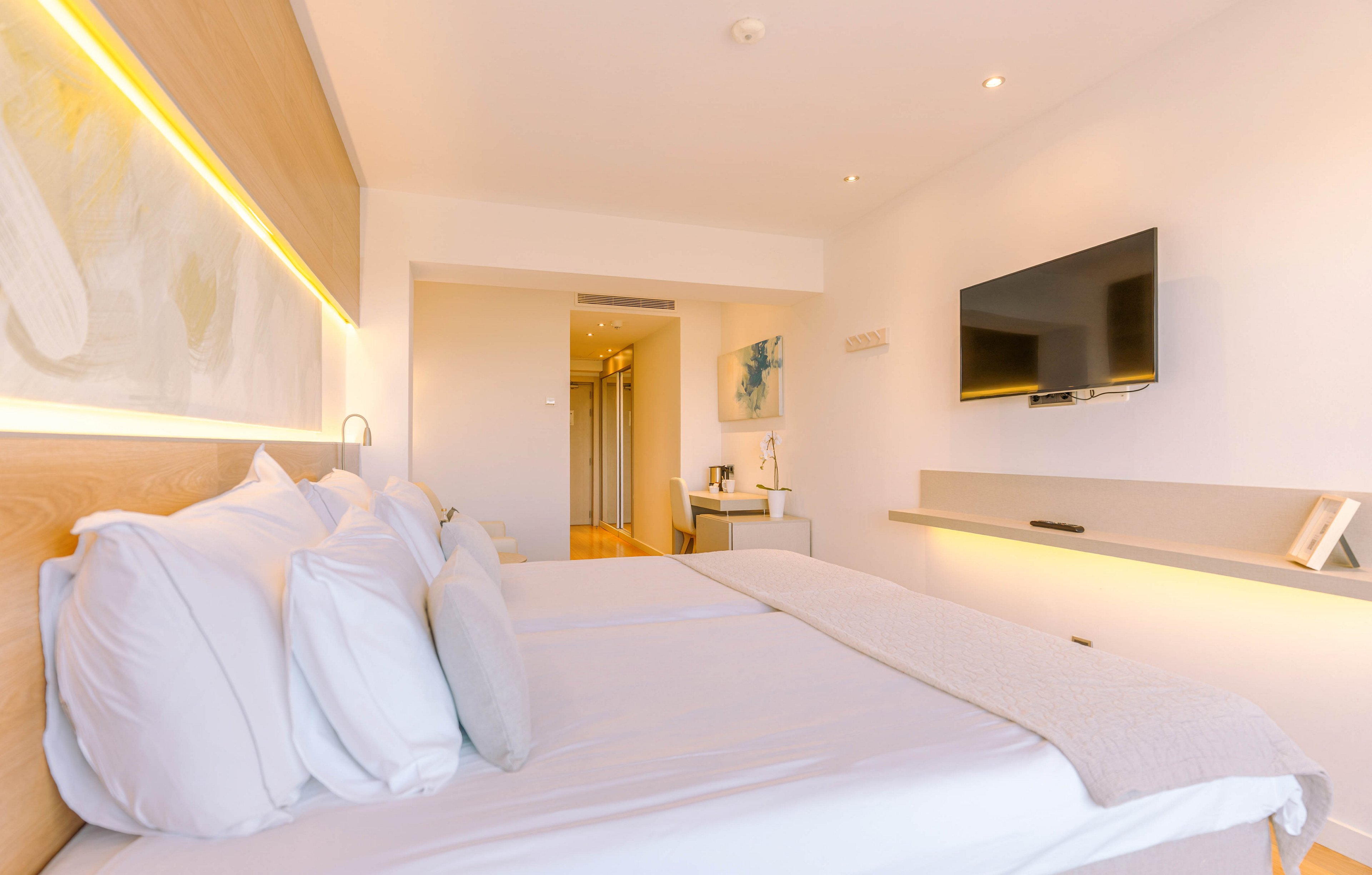 A neatly made bed with white linens dominates a bright modern hotel room. Warm recessed lighting runs along the headboard and a floating shelf, creating a cozy atmosphere. A flat-screen television is mounted on the wall opposite the bed, with a small desk area and artwork in the background. The room features minimalist decor, light wood accents, and a clean, uncluttered layout.