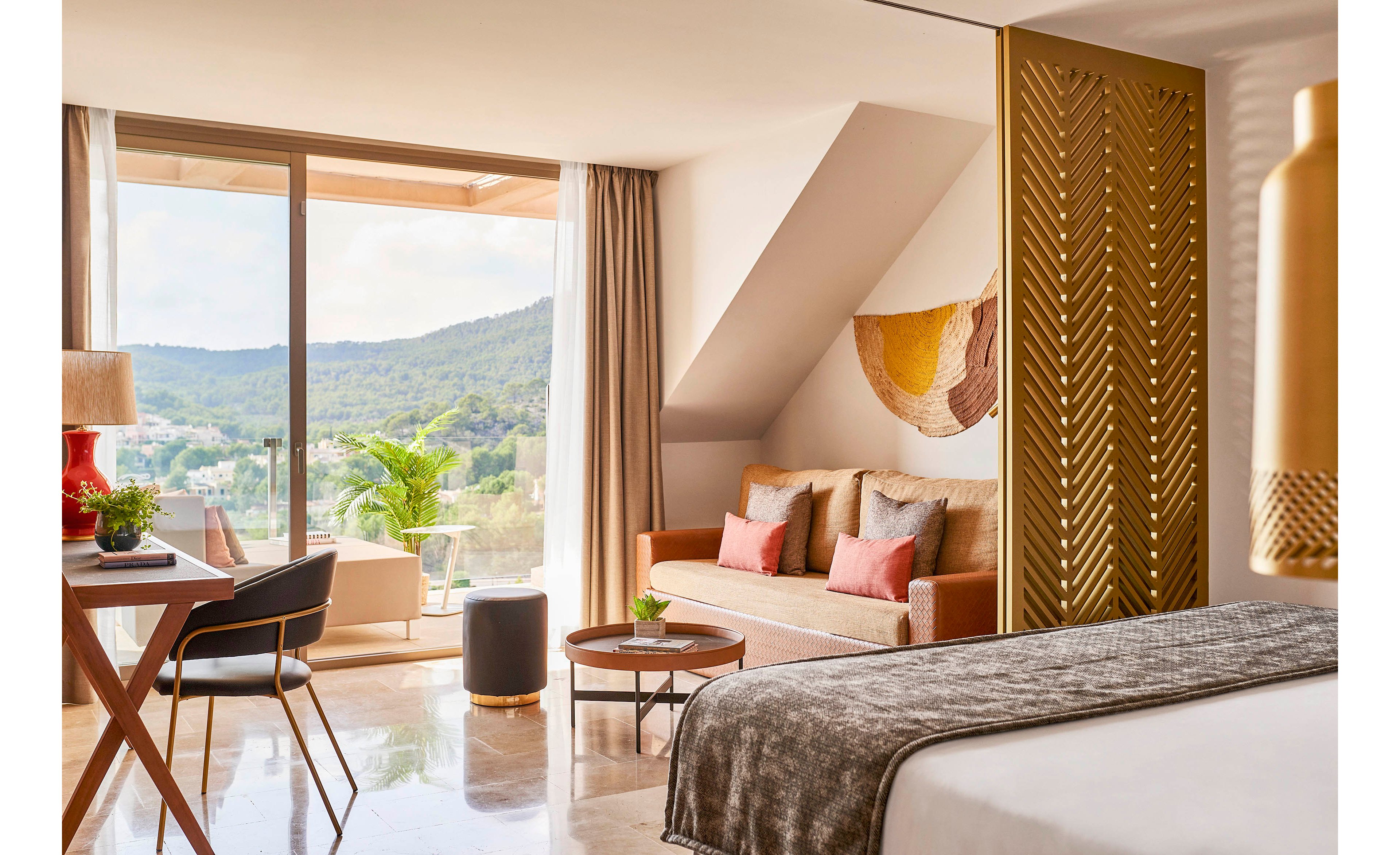 A contemporary hotel room features a neatly made bed, a cozy sofa with colorful cushions, and a small seating area. Large sliding glass doors open to a balcony overlooking green hills and a distant town. Warm neutral tones, gold accents, and soft textiles create a relaxed, upscale atmosphere. Natural light floods the space, highlighting plants, decorative wall art, and minimalist furniture.