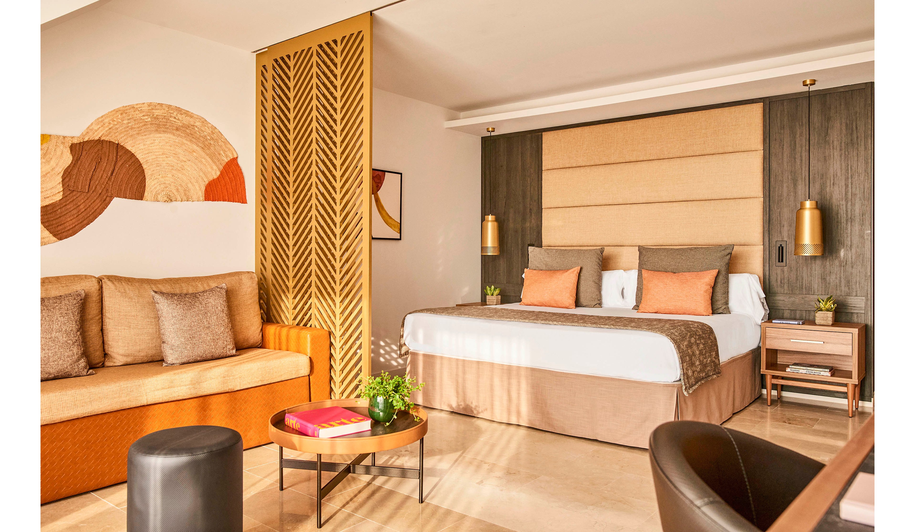 A contemporary hotel room features a large bed with neutral bedding and soft orange accent pillows. To the left, a beige sofa with cushions sits beneath a textured wall mural in earthy tones. A decorative wooden partition with geometric cutouts separates the sleeping area from the rest of the space. A round coffee table with books and a small plant, along with modern side tables and pendant lights, complete the cozy, inviting atmosphere.
