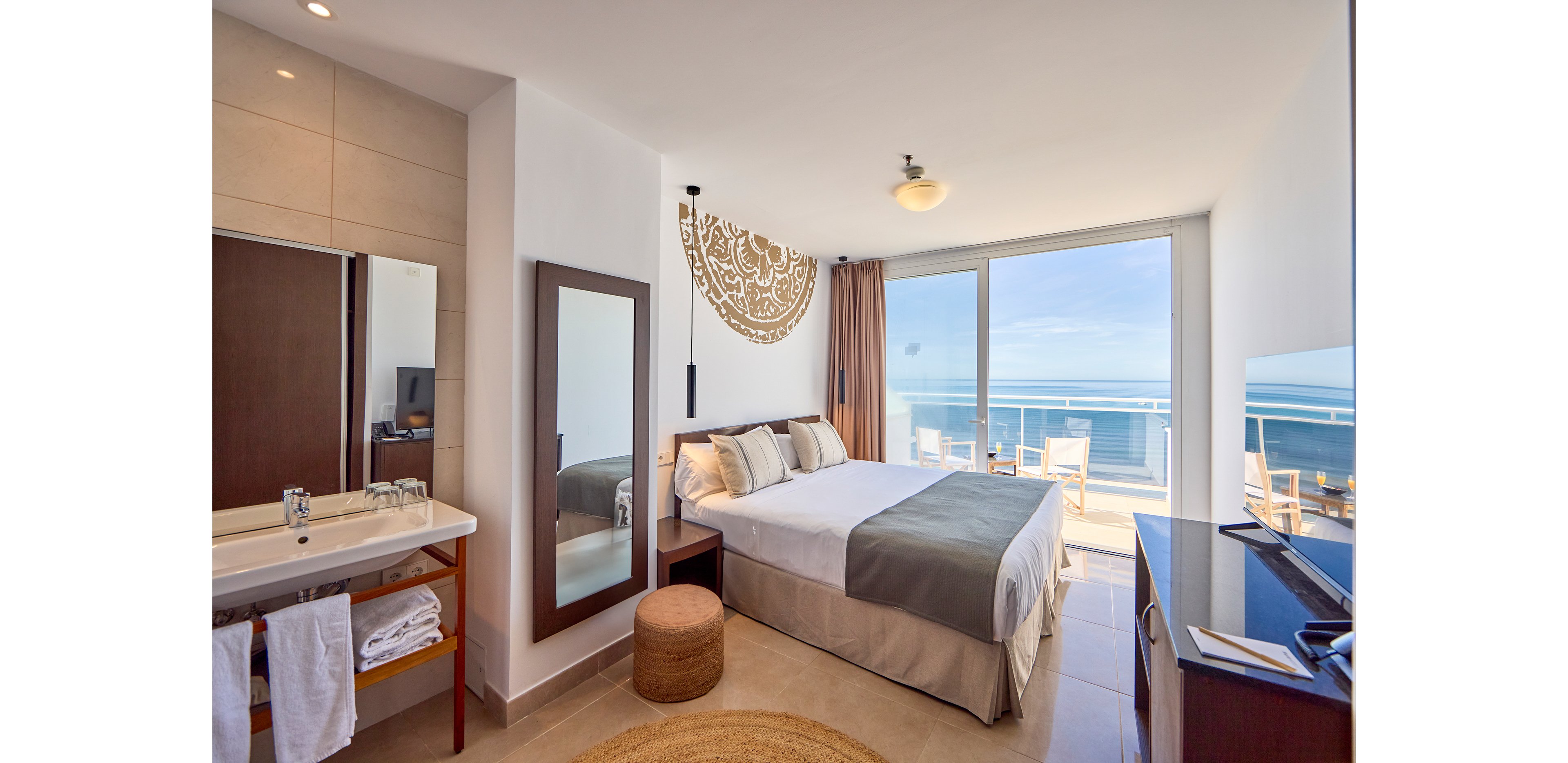 A contemporary hotel room featuring a comfortable double bed with neutral-toned bedding, a wall-mounted mirror, and a small seating area. The room opens to a balcony with a stunning ocean view under a clear blue sky. A minimalist decor style is complemented by natural light streaming through the glass doors.