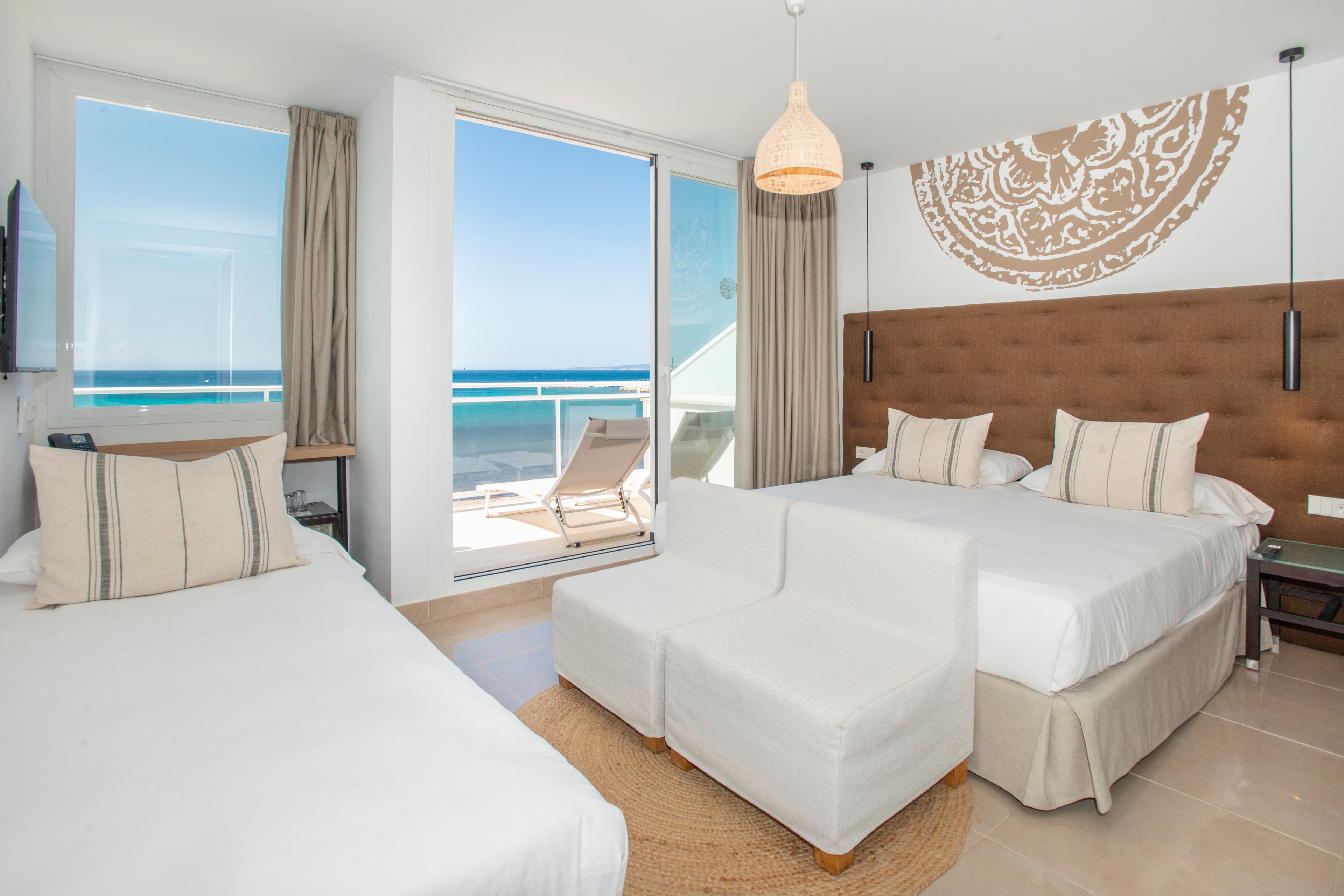 A spacious and bright hotel room featuring two beds and a seating area, overlooking a stunning ocean view through large glass doors. The room is decorated in neutral tones with a contemporary style, including a patterned wall accent and minimalist furniture. A private balcony with lounge chairs adds to the relaxing ambiance.