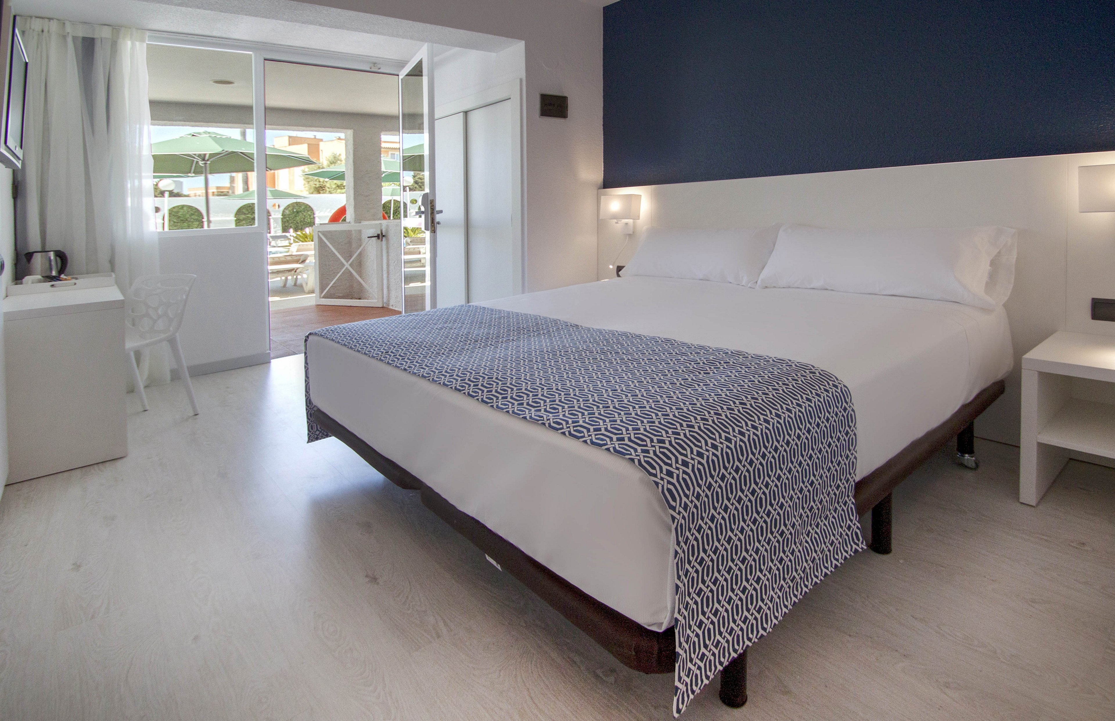 Contemporary hotel bedroom featuring a large double bed with white linens and a patterned blue throw. The room has light wood flooring, minimalist white furniture, and a deep blue accent wall behind the bed. Glass doors open to a sunlit terrace with outdoor seating and views of a pool area. Overall, the space feels clean, airy, and inviting for guests.